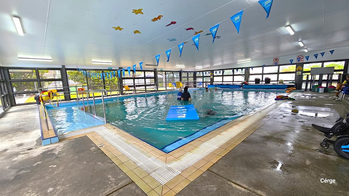 Right next to the tinted glass sliding doors with black frames is the Indoor program pool heated to 33 degrees Celsius and is restricted to rehabilitation and gentle exercise users over the age of 16 in Mudgeeraba Aquatic Centre. The Parent room inside has a wooden bench, a blue plastic picnic table, and a table with a colorful coral structural design. To the back is a glass window and a black door. The Parent room has a smooth cemented floor with a white ceiling.