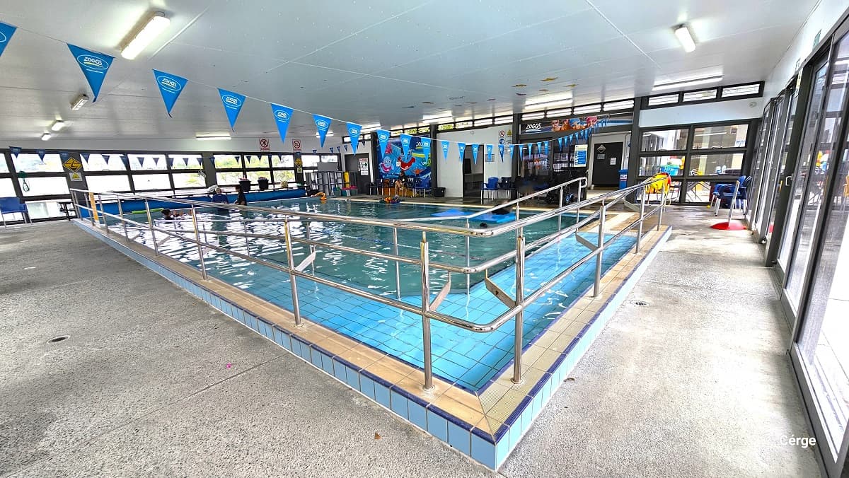 Ramp access with metal handrails in the indoor aquatic pool. Different seating options are accessible throughout the poolside area. From this position, there are two more doors next to the tinted glass sliding doors. On the wall between the two doors is a large poster with cute swimming pool graphics.