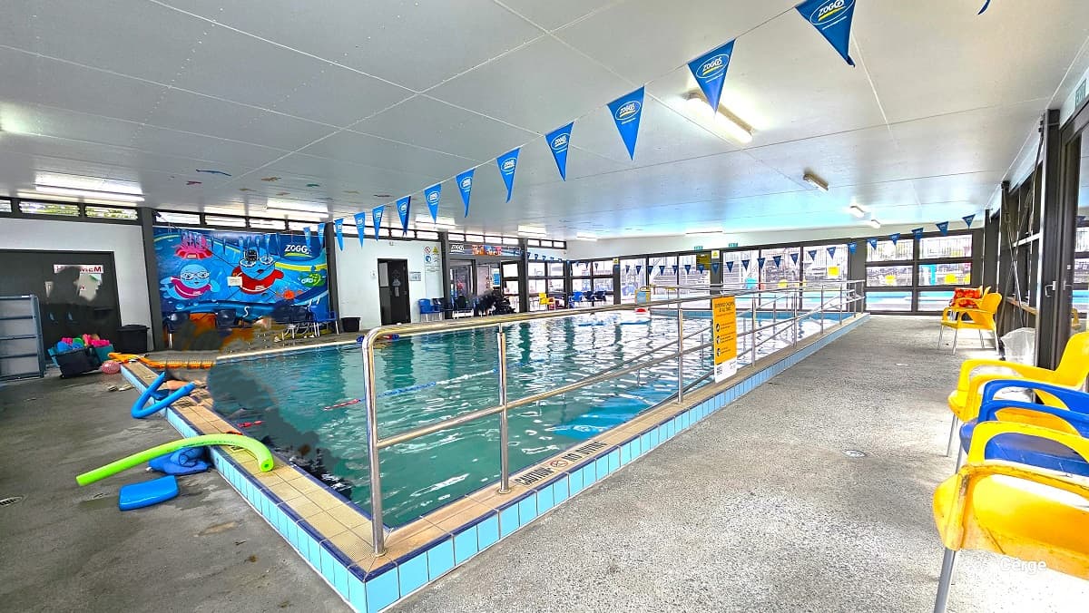 View opposite the wall with the cute swimming pool graphics. There are several pool floating toys on the poolside. A “Caution No Diving” sign is placed on the pool edge with metal handrails. Two strings of blue triangular flags across the pool are attached to the white ceiling. Blue and yellow chairs are available around the pool deck. The area is illuminated by artificial lighting and natural light from the tinted glass walls.