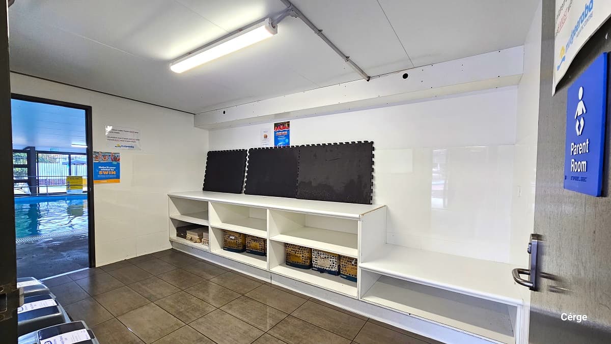 Parent room at Mudgeeraba Aquatic Centre. There are three grey garbage bins inside, a white cabinet, and three large black mats on top of the cabinet. On the lower space of the cabinet are net bags and books. The blue Parent Room sign can be found on the black door with a stainless steel door handle. The Parent room has a grey tiled floor, white walls, and ceiling.