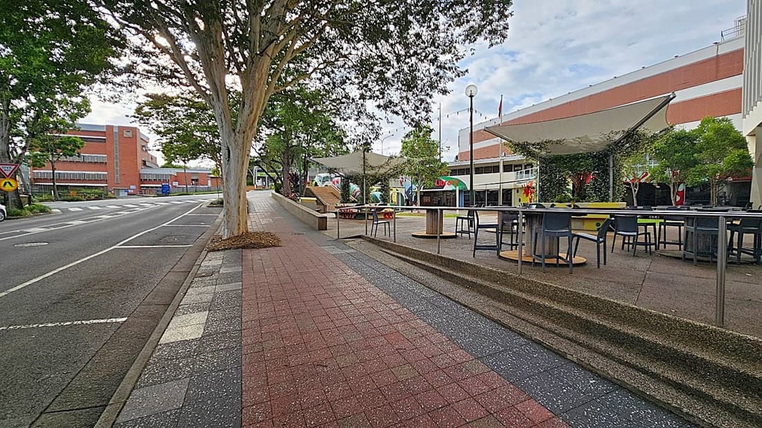 A street scene with a footpath on the left featuring a metal railing and a grey chair. Trees line the path, providing shade. On the right, a road with parked cars and a row of buildings, including a small structure with a red awning. Several bins are lined up against a building. The pavement is a mix of red and grey tiles, and a large tree trunk is visible in the foreground.