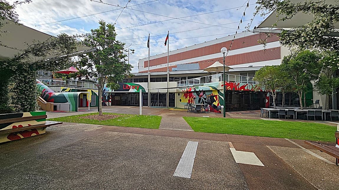 A courtyard with a library in the background. The library has colourful murals on its walls and a sign reading "Library" above the entrance. Two flagpoles stand in front of the building. On the left, a covered area with greenery and a bench. On the right, tables and chairs under trees. The ground is a mix of grass and paved paths, and string lights are strung overhead.
