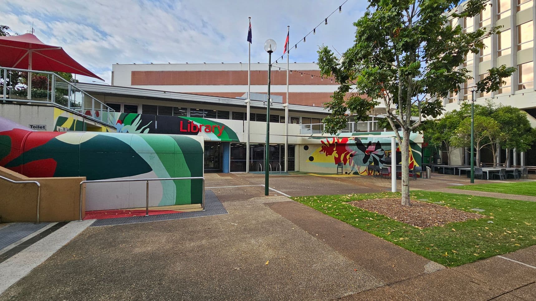 A library building with a colourful mural on the left side, featuring abstract shapes in green, red, and yellow. The entrance is in the centre, with the word "Library" in red. Three flagpoles stand in front, and a string of lights is strung above. On the right, there are trees and outdoor seating. The ground is a mix of concrete paths and grass patches. A red umbrella is visible on the left.