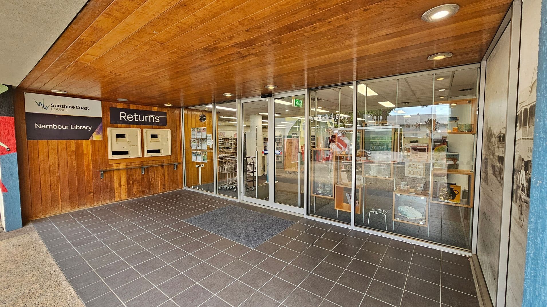 Entrance to Nambour Library with a wooden ceiling and tiled floor. On the left, there are two return slots under a "Returns" sign, and a "Sunshine Coast Council Nambour Library" sign. The glass doors in the centre lead into the library, revealing bookshelves and a cart inside. Posters and notices are displayed on the glass. On the right, a large window shows a historical photo and some library displays.