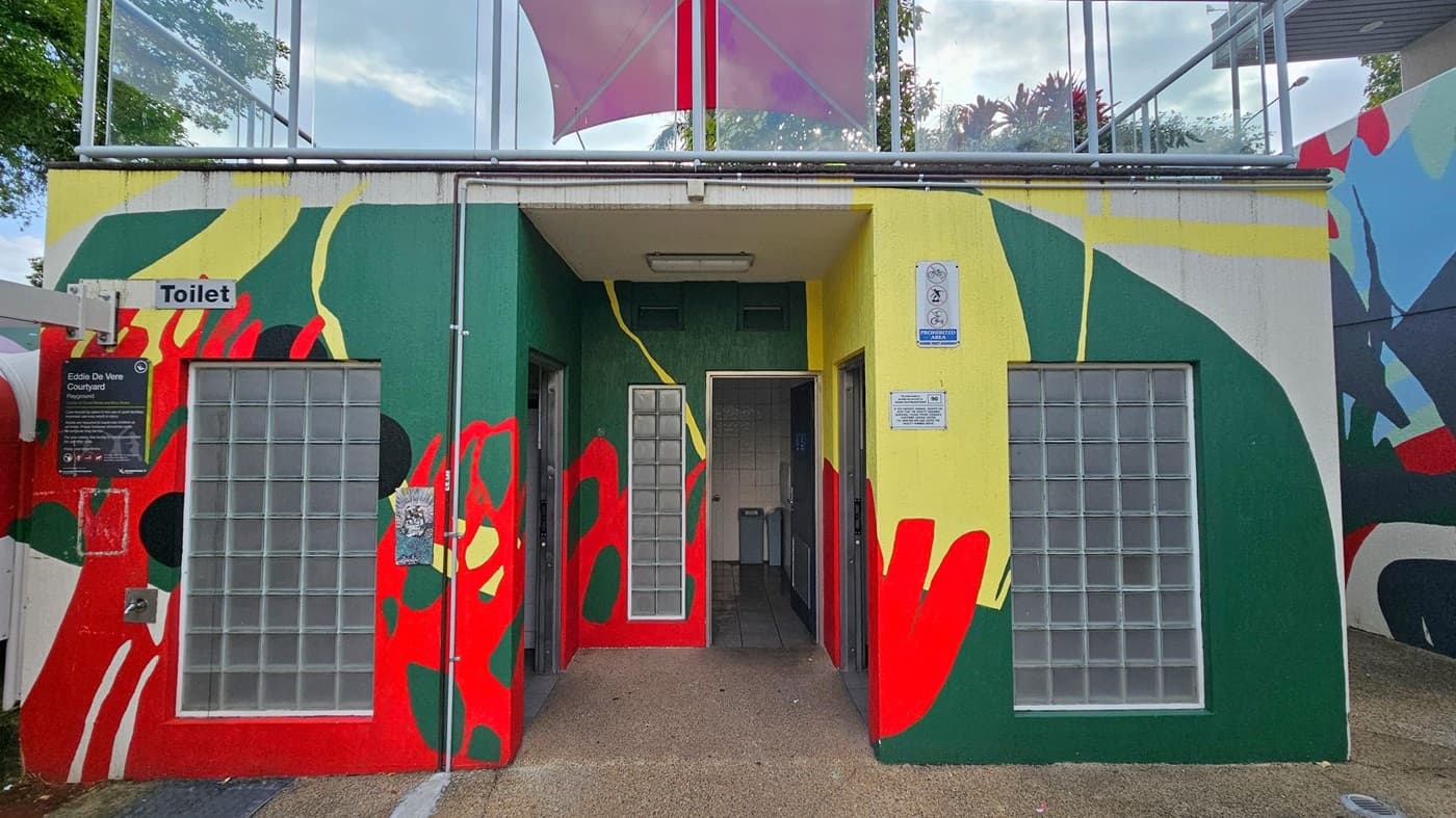 A public toilet building with a colourful mural featuring green, red, yellow, and black abstract shapes. The entrance is in the centre, flanked by glass block windows. A sign above the left side reads "Toilet," and there's a plaque for "Eddie De Vere Courtyard" below it. The ground is paved, and a pink canopy is visible above. Trees and a railing are in the background.
