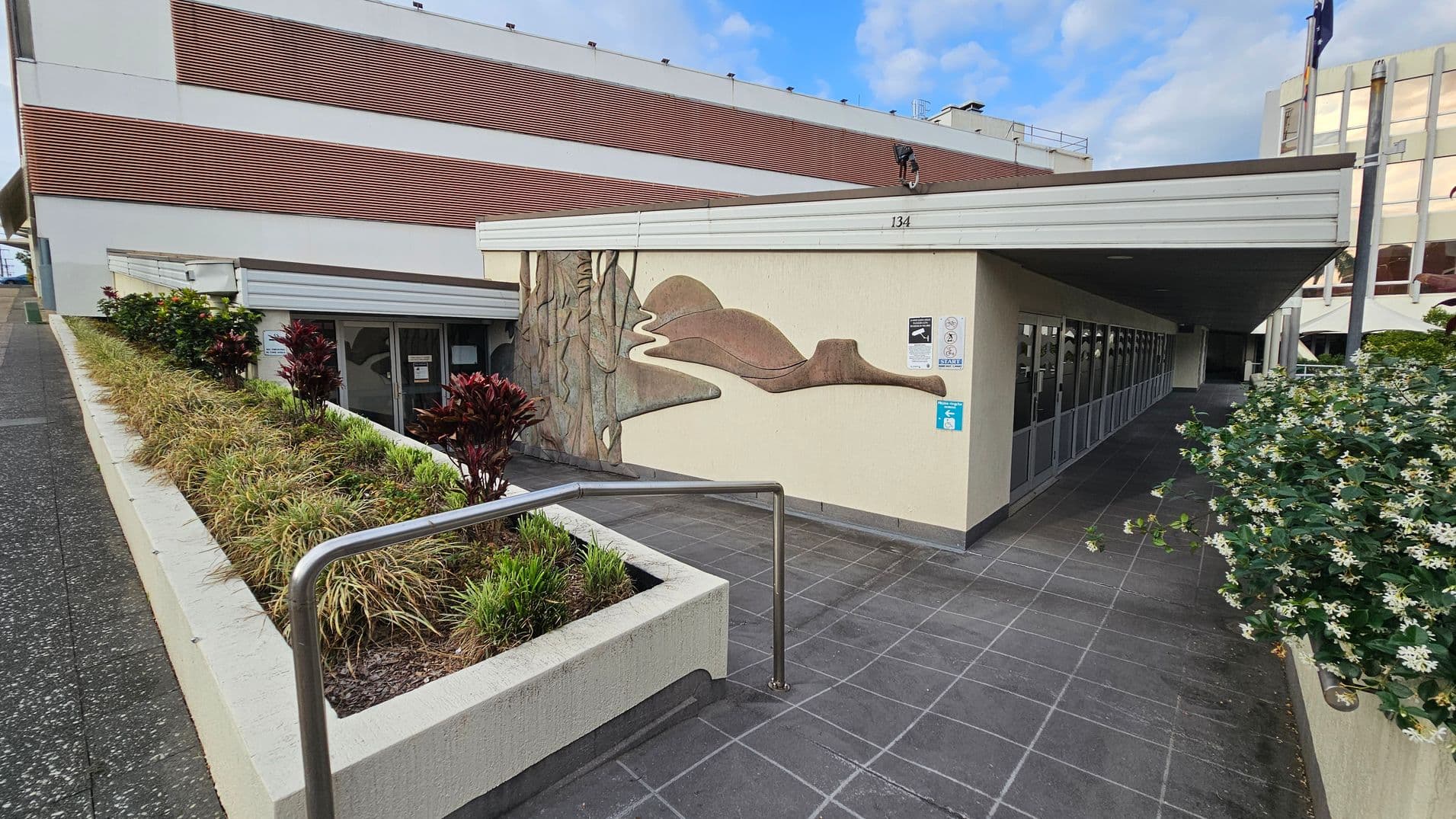 A building with a mural on the wall, featuring abstract shapes in earthy tones. On the left, there's a sloped path with a metal railing and a planter box filled with grass and red-leaved plants. The right side shows a row of glass doors under a flat roof. In the foreground, a bush with white flowers is visible. The ground is paved with dark tiles, and the building has a mix of white and brown exterior panels.