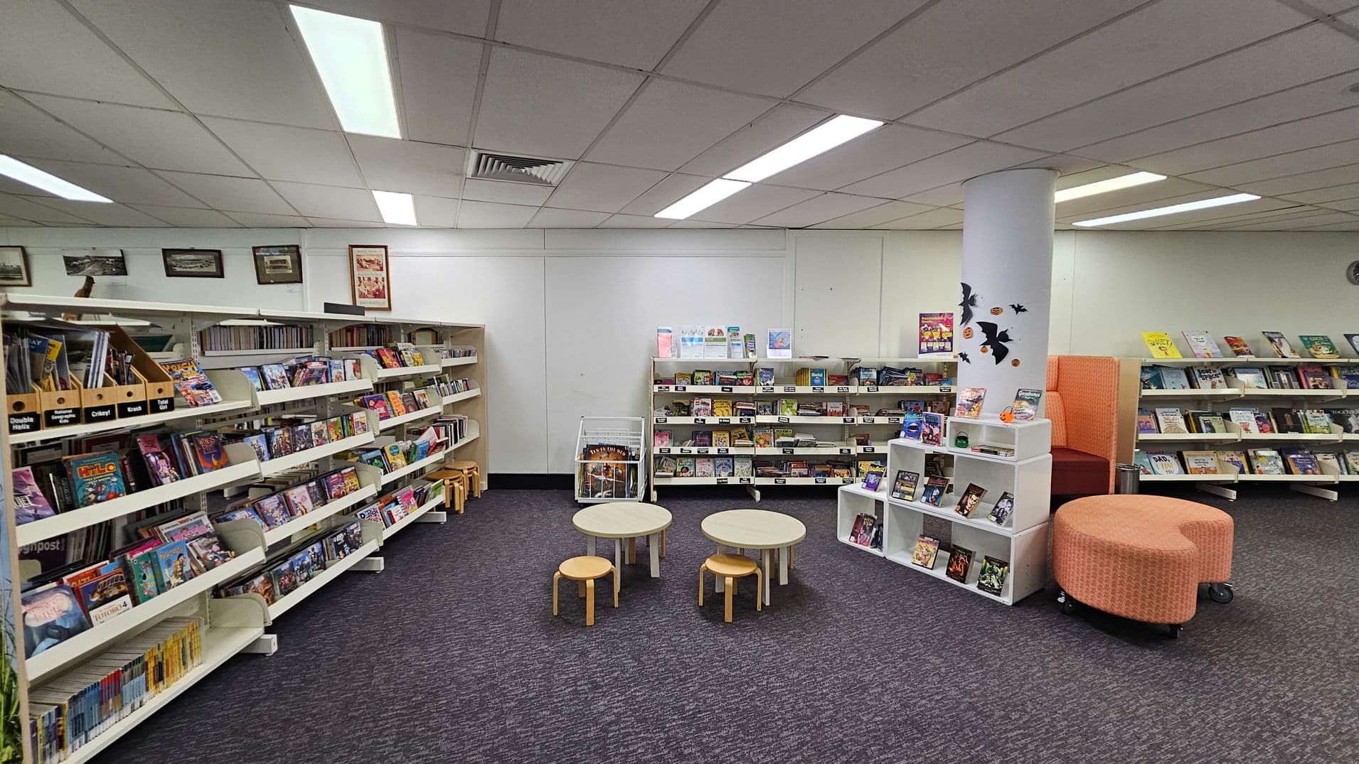 A library interior with bookshelves lining the left and right walls, filled with various books and magazines. In the centre, there are small round tables with wooden stools. A white pillar decorated with black bat cutouts stands near an orange cushioned seating area. The floor is carpeted in dark grey, and the ceiling has fluorescent lights and a grid pattern.