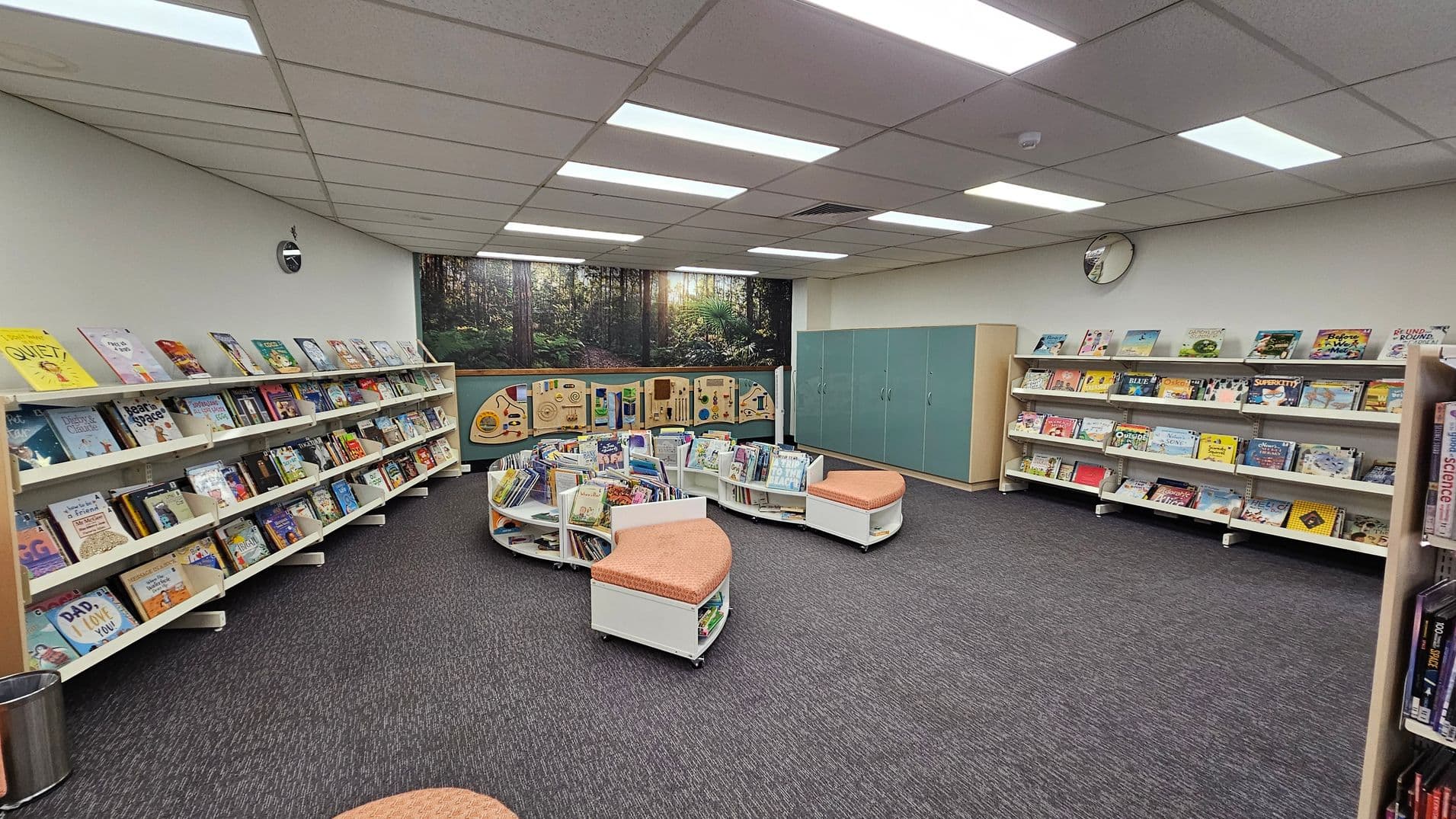 A library room with bookshelves lining the left and right walls, filled with children's books. In the centre, there are curved shelves with more books and cushioned seating. The back wall features a mural of a forest scene and green cabinets. The ceiling has fluorescent lights, and the floor is carpeted in dark grey. A round clock is mounted on the right wall.