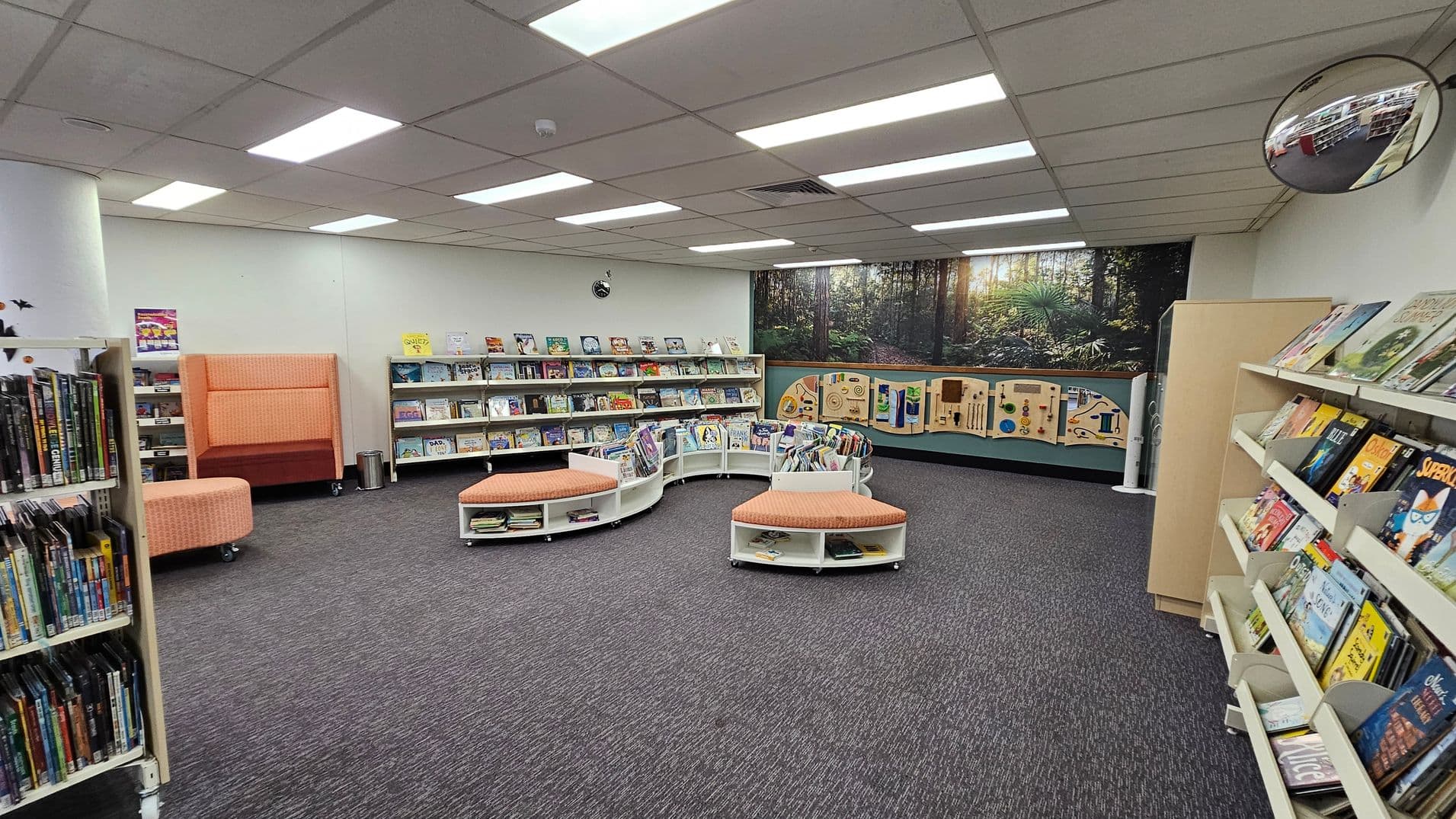 A library interior with grey carpeted flooring and a ceiling with fluorescent lights. On the left, there are shelves filled with books and DVDs. In the centre, there are curved bookshelves with orange cushioned seating. The back wall features a large mural of a forest scene and interactive panels. On the right, more bookshelves display children's books. A circular mirror is mounted on the upper right wall.