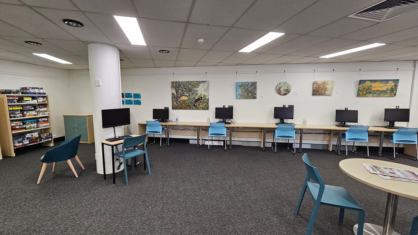 A library interior with a grey carpeted floor and a white panelled ceiling with fluorescent lights. On the left, there are bookshelves labelled "Audiobooks" filled with items. A teal chair is positioned near a small table. On the right, there are computer stations with blue chairs and monitors on wooden desks. A large painting hangs above the computers. A white column stands in the centre of the room.