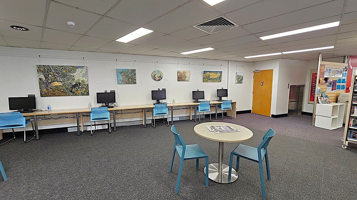 A room with a row of computers on desks against the left wall, each accompanied by a blue chair. Above the computers, several colourful paintings are displayed. In the foreground, a round table with two blue chairs has a newspaper on it. The floor is carpeted in grey, and the ceiling has fluorescent lights. On the right, there is a wooden door and a small shelving unit with books and pamphlets.