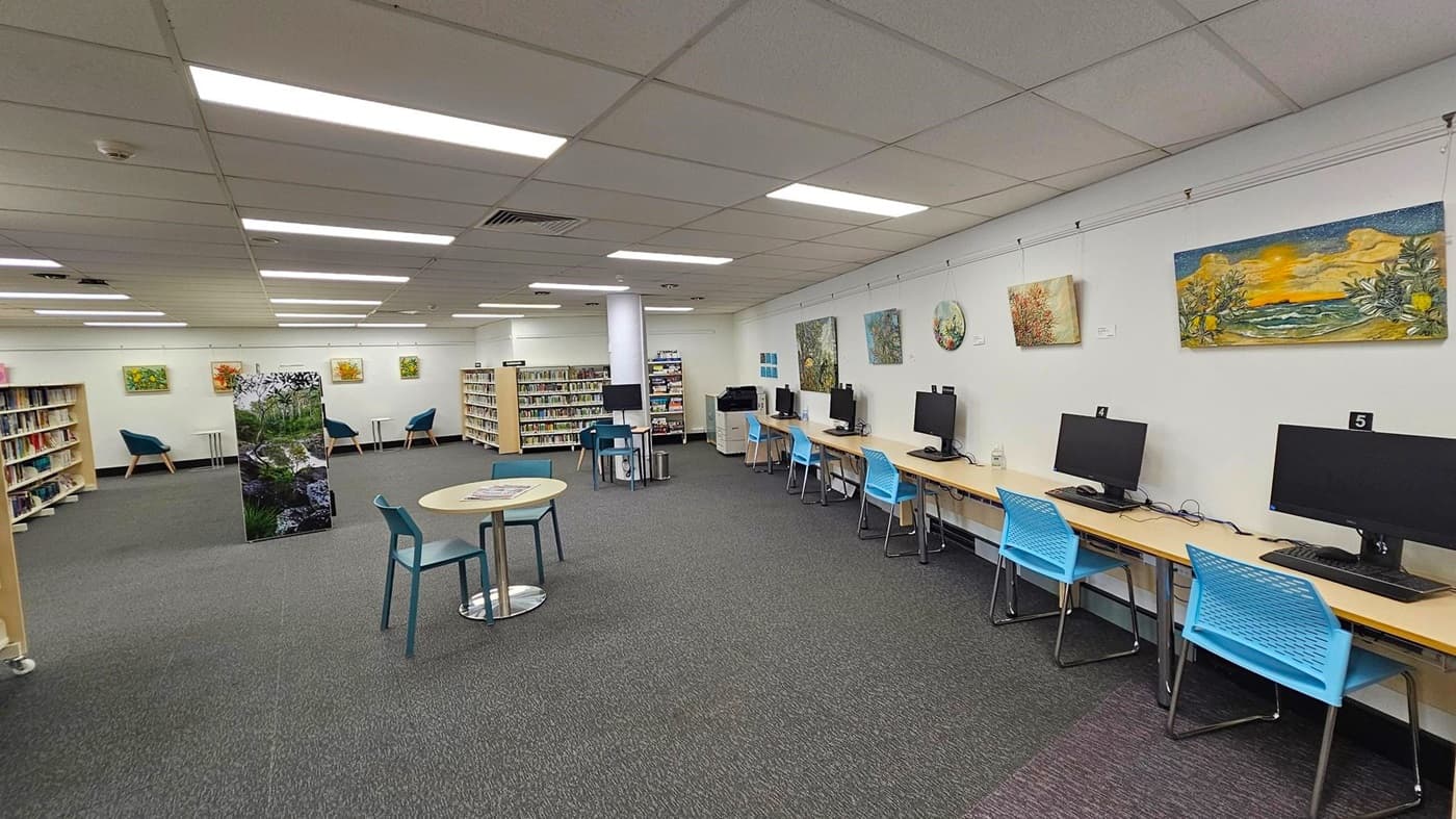 A library interior with a row of computer stations on the right, each with a monitor and a blue chair. The left side features bookshelves filled with books and a few teal armchairs. In the centre, there are small round tables with chairs. The walls display various paintings, and the ceiling has fluorescent lighting. The floor is carpeted in grey.