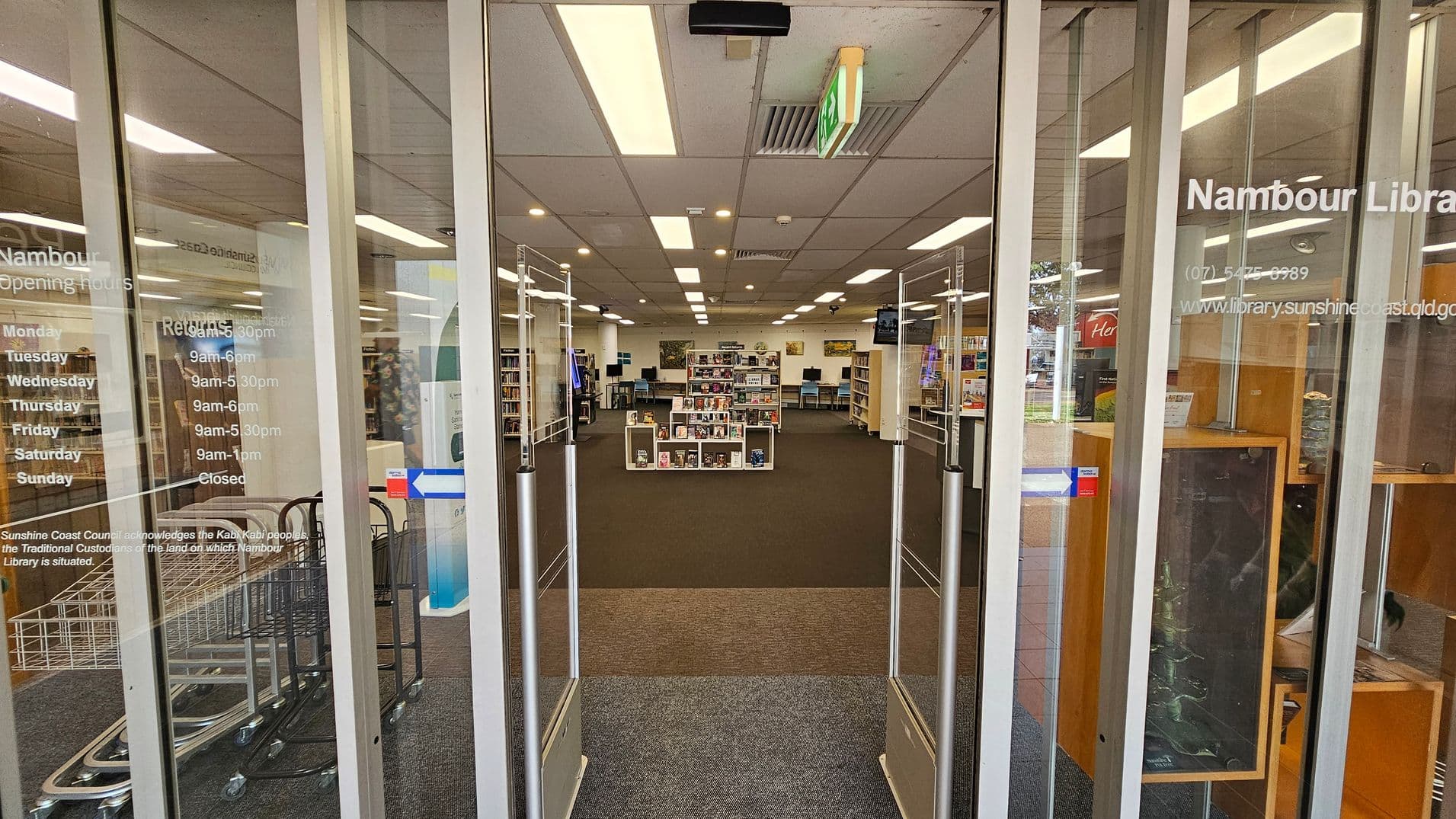 Glass entrance doors to a library with opening hours listed on the left. Inside, the library has a carpeted floor and a grid of fluorescent lights on the ceiling. In the centre, there are bookshelves filled with books and media. To the right, there is a wooden display case. In the background, more shelves and computer stations are visible. An exit sign hangs from the ceiling.