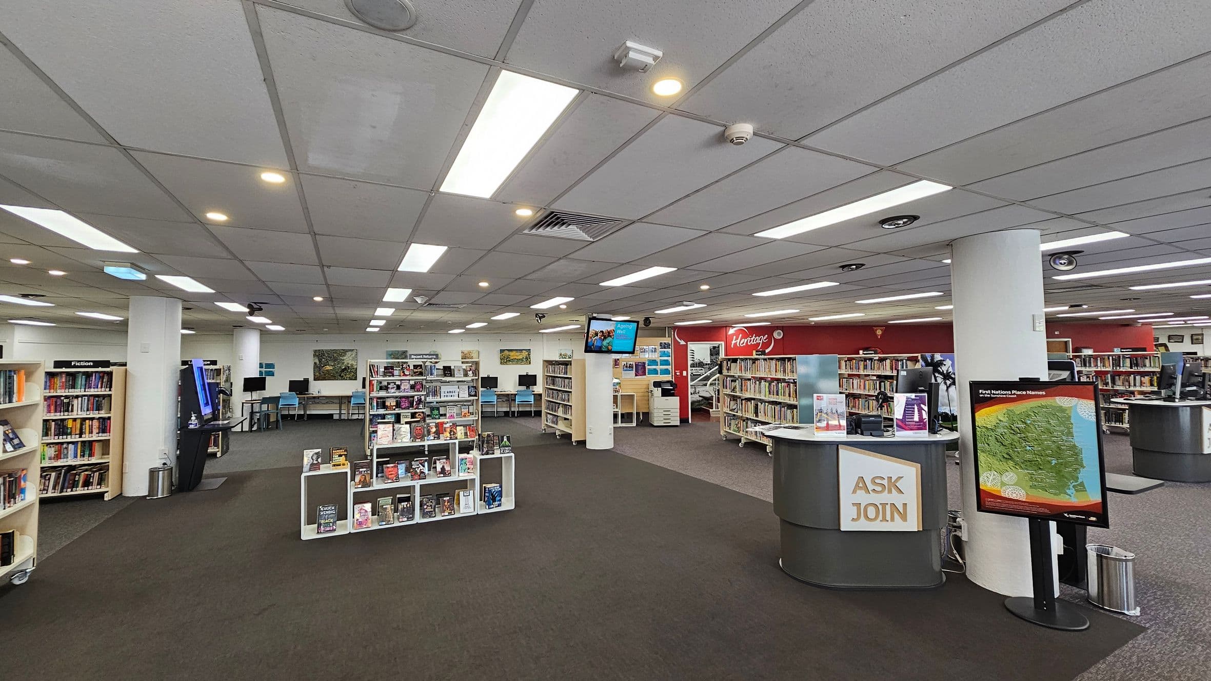 A spacious library interior with a grey-tiled ceiling and bright fluorescent lights. In the foreground, a circular desk with a sign reading "ASK JOIN" and a map display. To the left, bookshelves labelled "Fiction" and a digital kiosk. In the centre, more bookshelves and a TV screen mounted on a pillar. The background features computer stations with blue chairs and a red wall with "Heritage" signage. The floor is carpeted in dark green and grey.