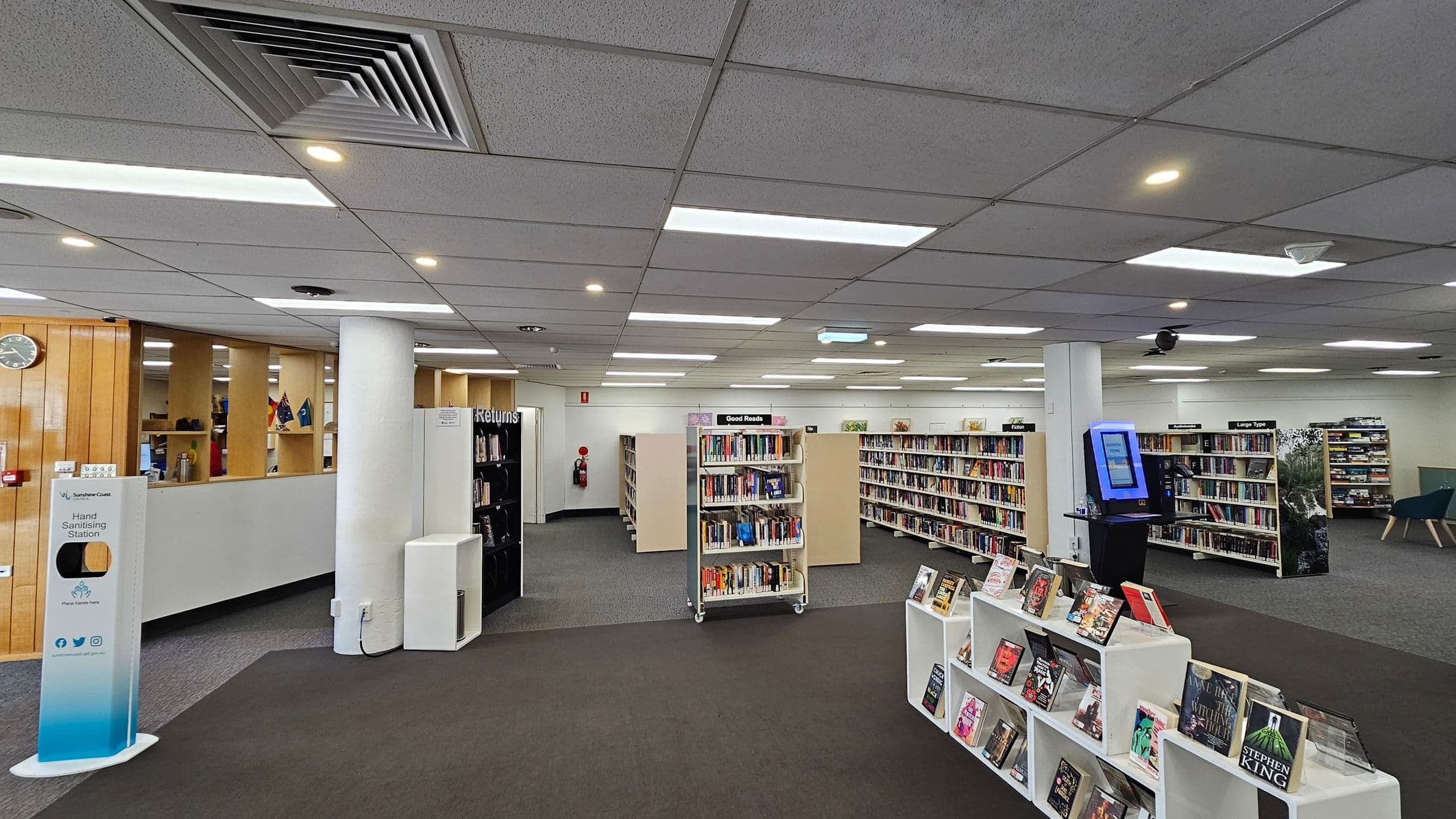 A library interior with a grey carpeted floor and a ceiling with recessed lighting and air vents. In the foreground, there are white shelves displaying books. In the middle, several bookcases are filled with books, labelled "Good Reads" and "Large Type". On the right, a digital kiosk stands near a column. On the left, a hand sanitising station is positioned near a wooden partition with a clock and small flags. A "Returns" section is visible in the background.