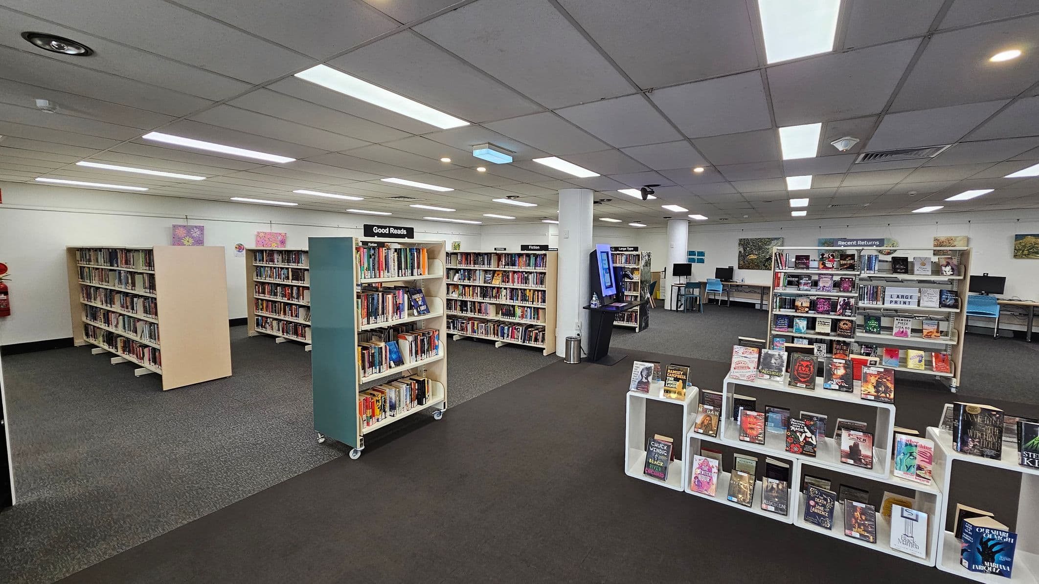 A library interior with several bookshelves filled with books. On the left, there are shelves labelled "Good Reads" and "Fiction". In the centre, a display features books on white stands. To the right, a "Recent Returns" section is visible. The background shows computer stations along the wall. The floor is carpeted in grey, and the ceiling has fluorescent lighting.