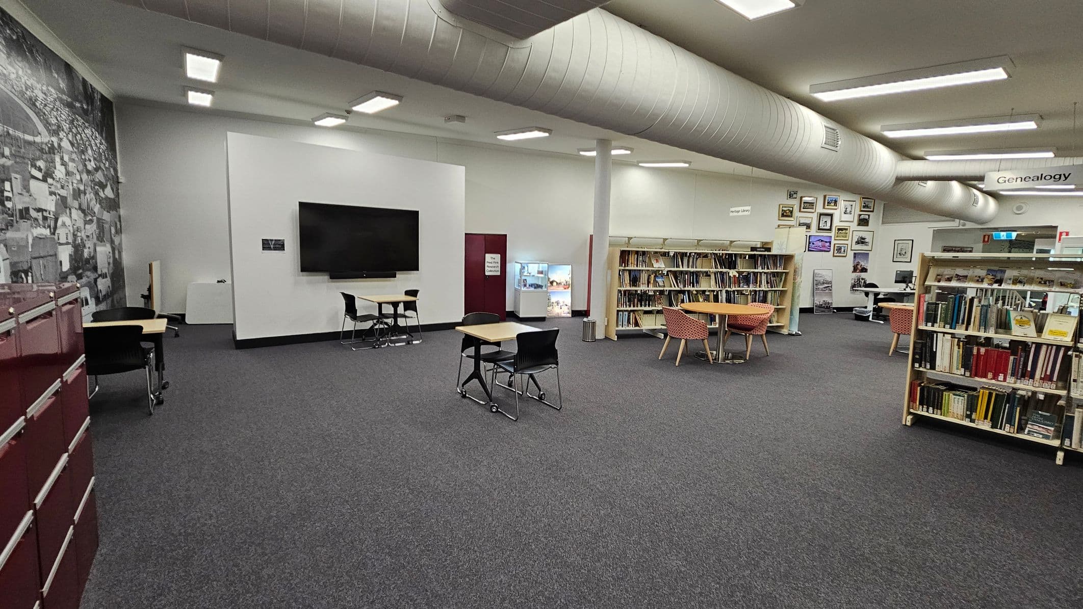 A room with a red wall featuring the word "Heritage" and arrows pointing towards an open doorway. To the left, there's a bulletin board with various posters and a printer below it. In the background, several computer workstations with blue chairs are lined up against a white wall. The floor is carpeted, and the ceiling has fluorescent lights. A small table with two blue chairs is on the right.