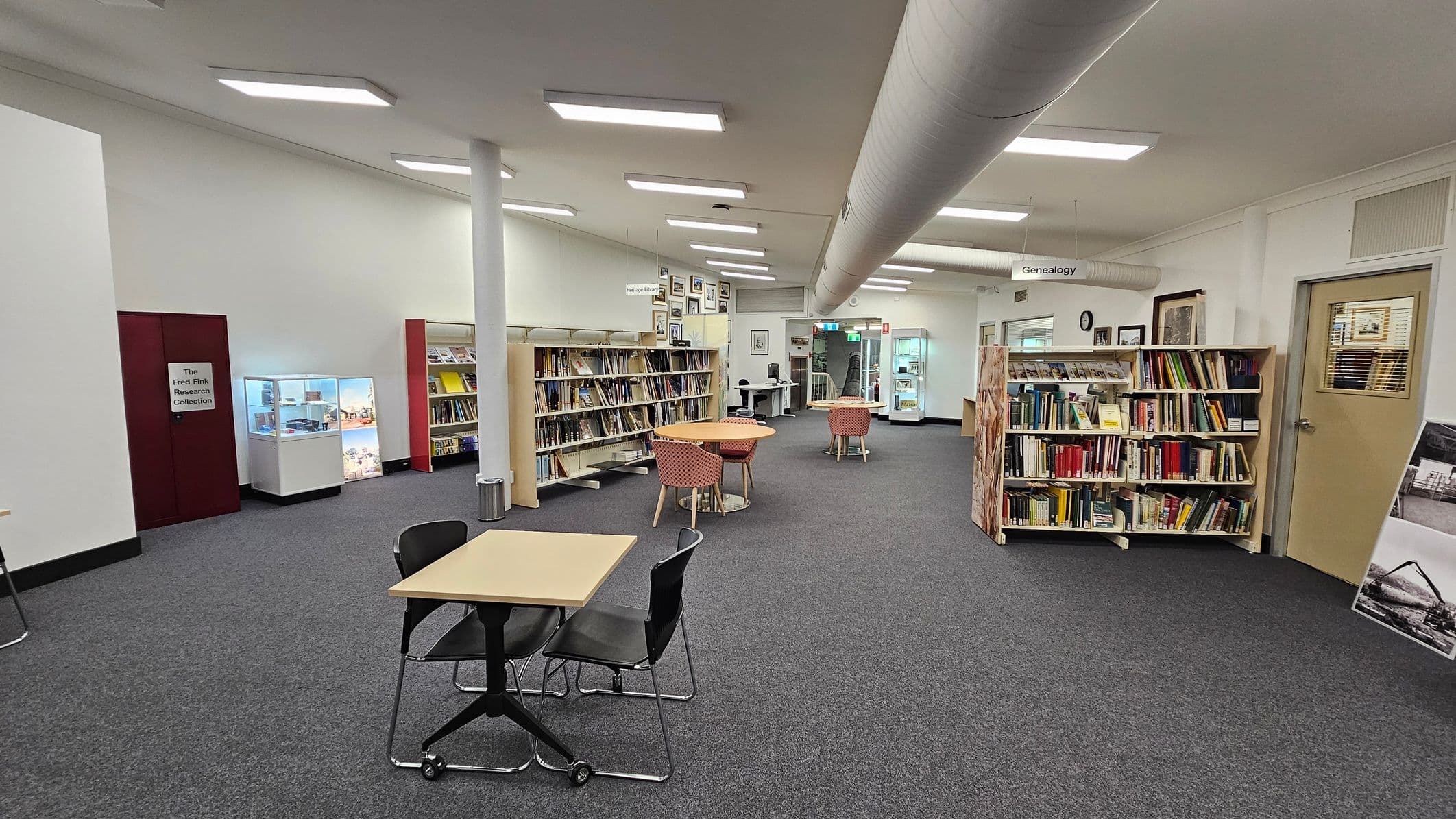A library interior with grey carpeted flooring. On the left, there is a display case and a section labelled "The Fred Fink Research Collection." In the centre, there are bookshelves filled with books and a few round tables with red-patterned chairs. On the right, more bookshelves are visible, along with a door. The ceiling has fluorescent lights and a large ventilation duct running across. In the background, there are more tables and a computer station.