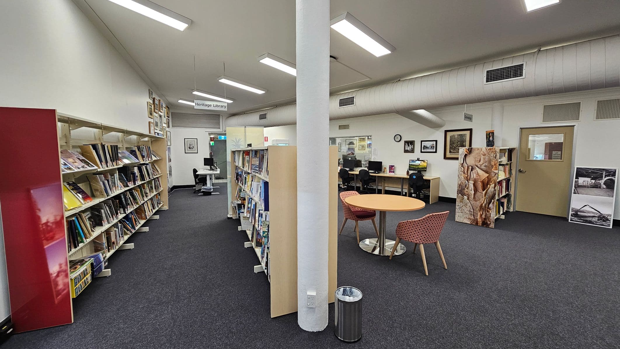 A library interior with a central white pillar. On the left, there are bookshelves filled with books and a red panel. In the middle, more bookshelves are visible. On the right, there's a round table with two chairs, a computer workstation with chairs, and framed pictures on the wall. A door with a window is also on the right. The floor is carpeted, and the ceiling has fluorescent lights and a large air duct. A small bin is near the pillar.