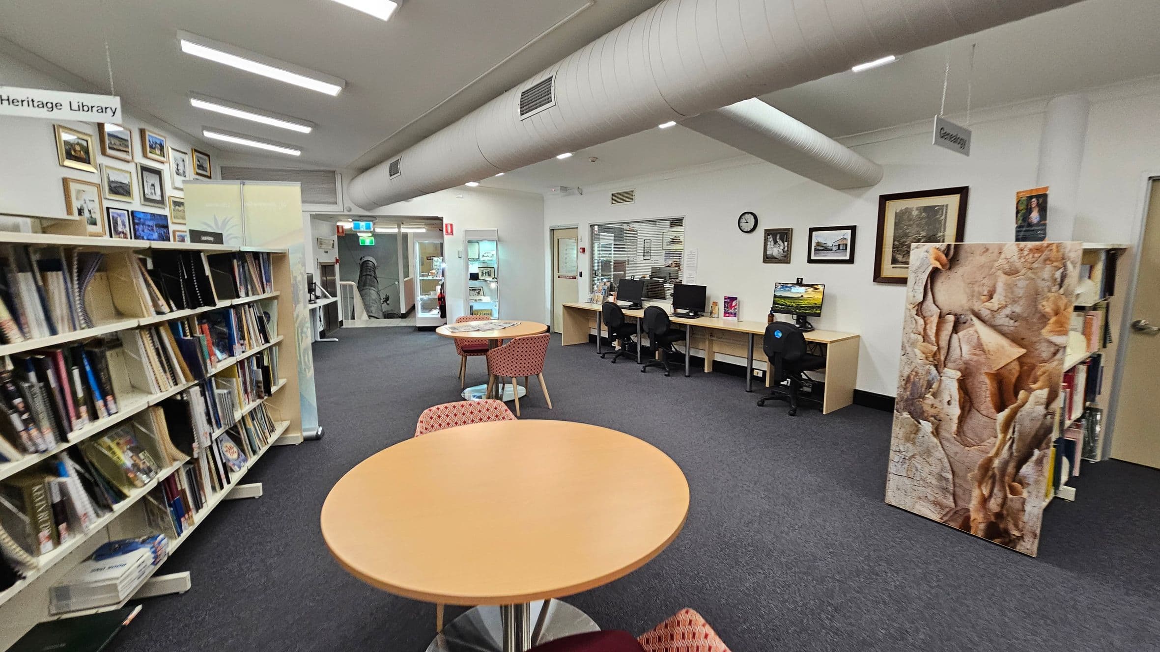 A library interior with bookshelves on the left filled with books and framed pictures above. In the centre, there are round tables with red-patterned chairs. On the right, a row of computer desks with chairs and a clock on the wall. A large artwork stands near the bookshelves. The floor is carpeted, and the ceiling has exposed ductwork and fluorescent lights. A sign reads "Heritage Library" on the left and "Genealogy" on the right.