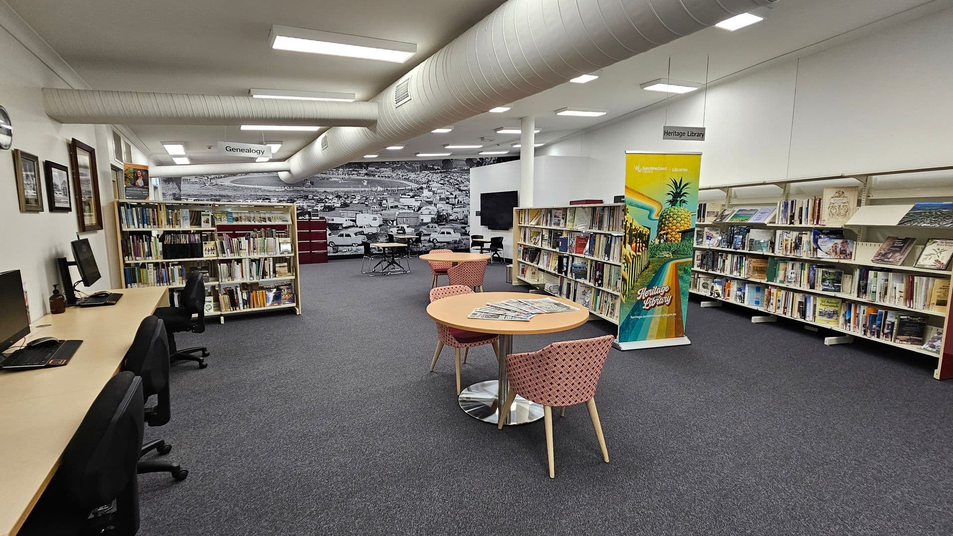 A library interior with a grey carpeted floor. On the left, there are computer desks with chairs and framed pictures on the wall. In the centre, round tables with patterned chairs hold newspapers. Bookshelves line the walls, filled with books. A colourful banner stands near the shelves, promoting the Heritage Library. The ceiling has exposed ductwork and fluorescent lighting. A large black-and-white mural of a townscape is visible in the background.