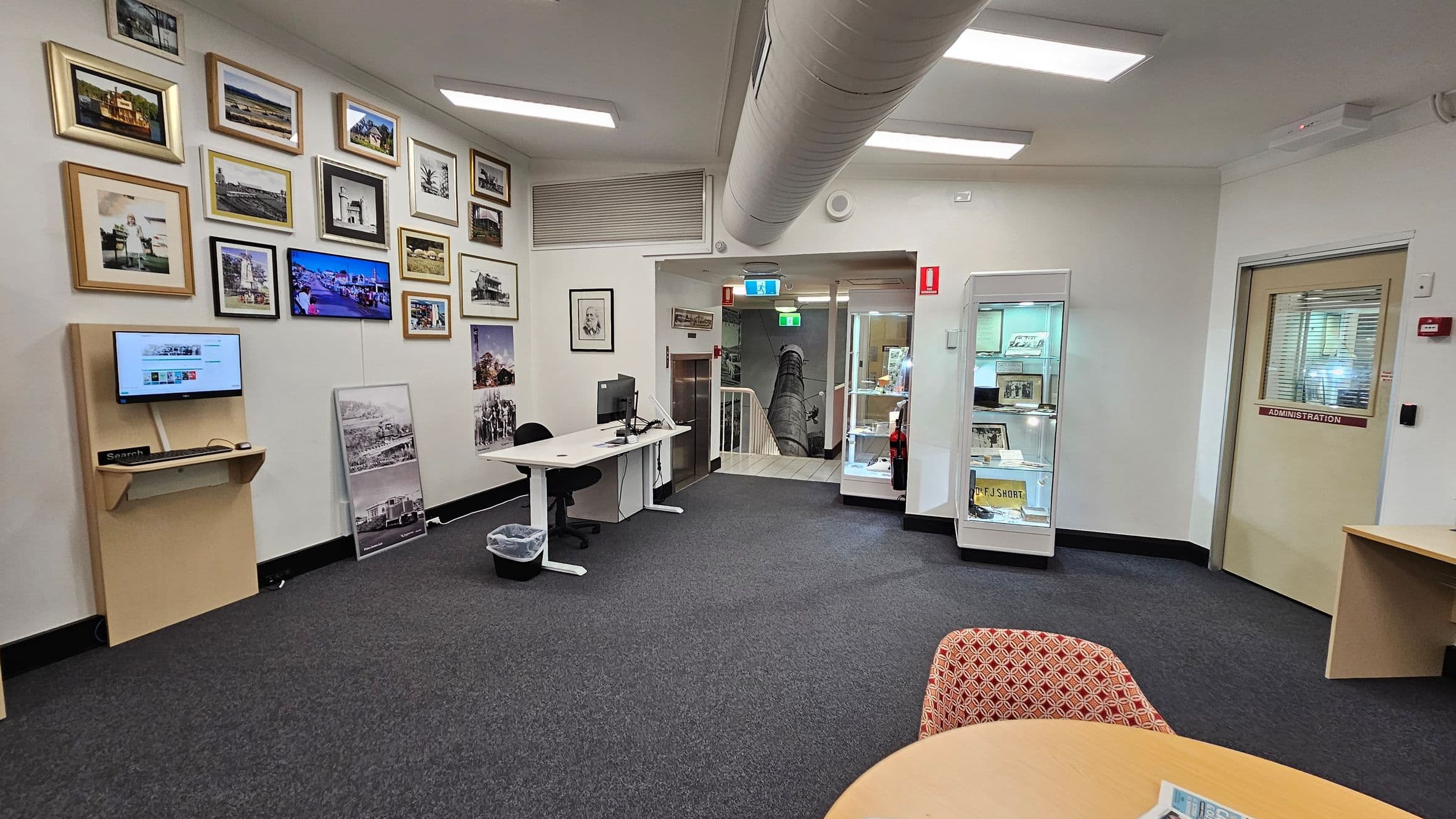A room with a grey carpet and white walls. On the left, a wall displays numerous framed photographs and a small monitor on a wooden shelf. Below, a desk with a computer and chair. In the centre, a doorway leads to a staircase, with a glass display cabinet nearby. On the right, a door marked "Administration" and a round table with a patterned chair. The ceiling has fluorescent lights and a large air duct.