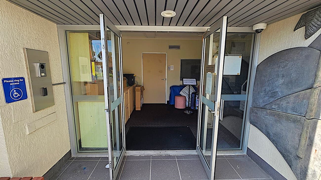 Glass double doors are open, leading into a building. On the left, there's a wall-mounted intercom system with a blue sign indicating wheelchair access. Inside, a beige door is visible at the back, with a small table and some items to the left. On the right, a decorative wall feature is partially visible. The ceiling is lined with grey panels, and a security camera is mounted near the entrance. The floor is tiled in dark grey.