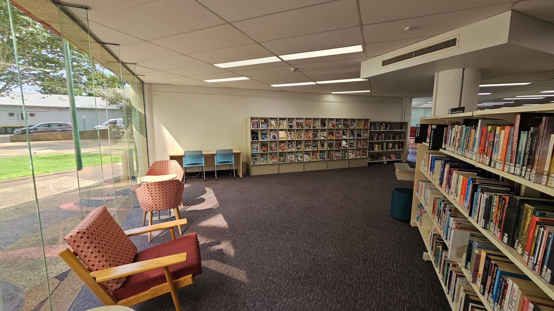 A library interior with a large glass window on the left, showing a view of the outside with grass and a road. Inside, there are two cushioned chairs and a small table near the window. Further back, two blue chairs are placed at a desk against the wall. The wall features shelves filled with magazines. On the right, there are bookshelves filled with books. The floor is carpeted in a dark colour, and the ceiling has rectangular lights.