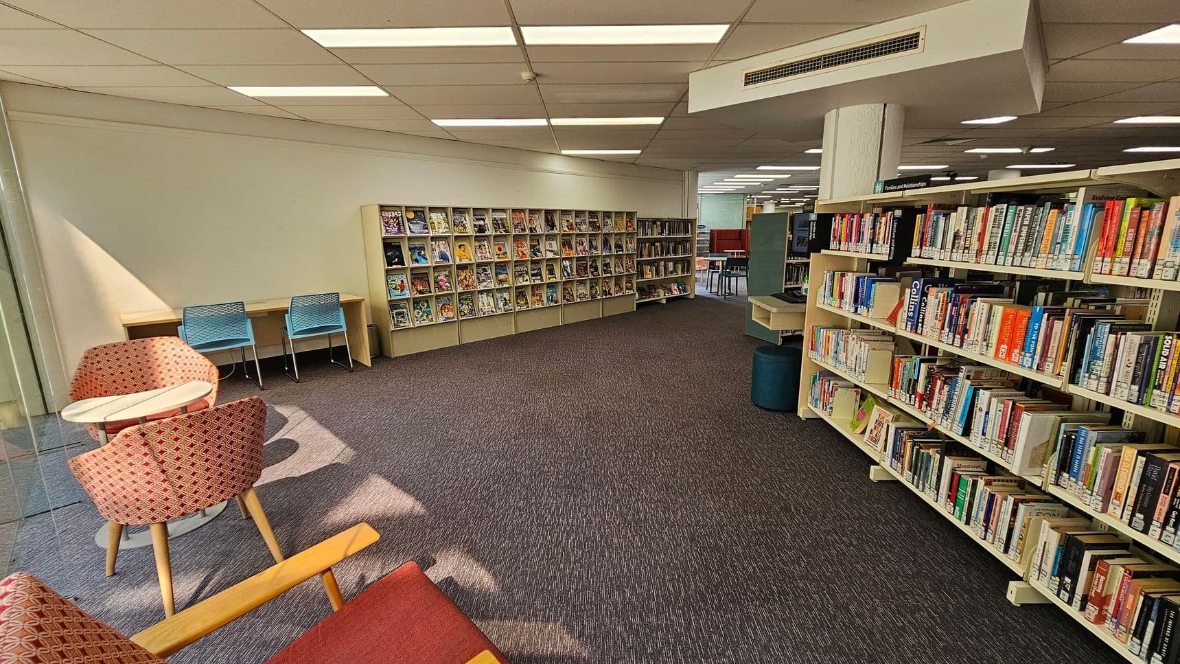 A library interior with bookshelves on the right filled with various books. In the middle, there are more shelves displaying magazines or comics. On the left, there are two blue chairs at a wooden desk and two red patterned chairs around a small round table. The floor is carpeted in a dark colour, and the ceiling has white panels with fluorescent lights.