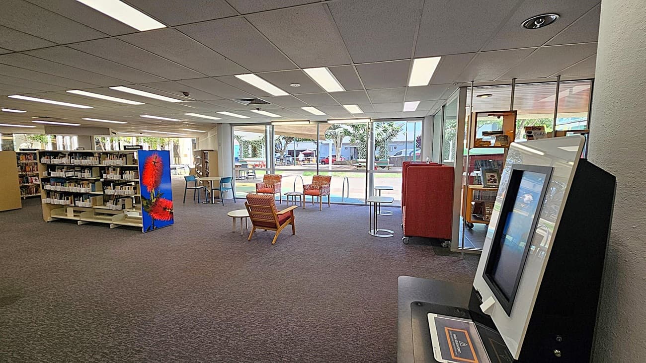 A library interior with a self-service kiosk on the right. In the centre, there are two orange chairs and a small round table on a carpeted floor. To the left, bookshelves are filled with books, and one end of a shelf features a large image of red flowers. Large windows in the background let in natural light, revealing an outdoor area with trees and a few people. The ceiling is lined with fluorescent lights.