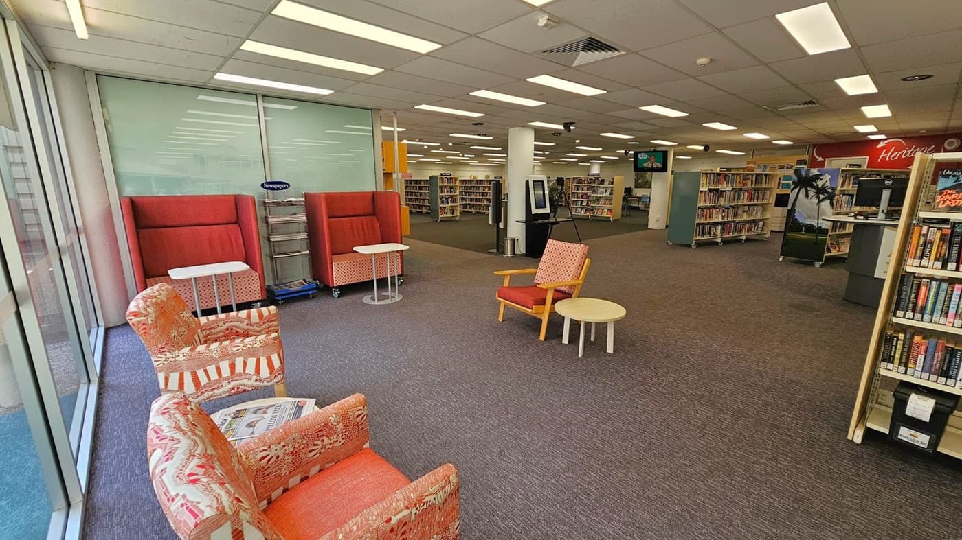 A library interior with a seating area on the left featuring patterned armchairs and small tables. Behind them are high-backed red chairs with additional tables. The floor is carpeted in a mix of grey and brown. In the centre, there are bookshelves filled with books, and a digital kiosk is visible. The ceiling has fluorescent lighting. On the right, more bookshelves and a computer station are present, with a sign reading "Heritage" in the background.