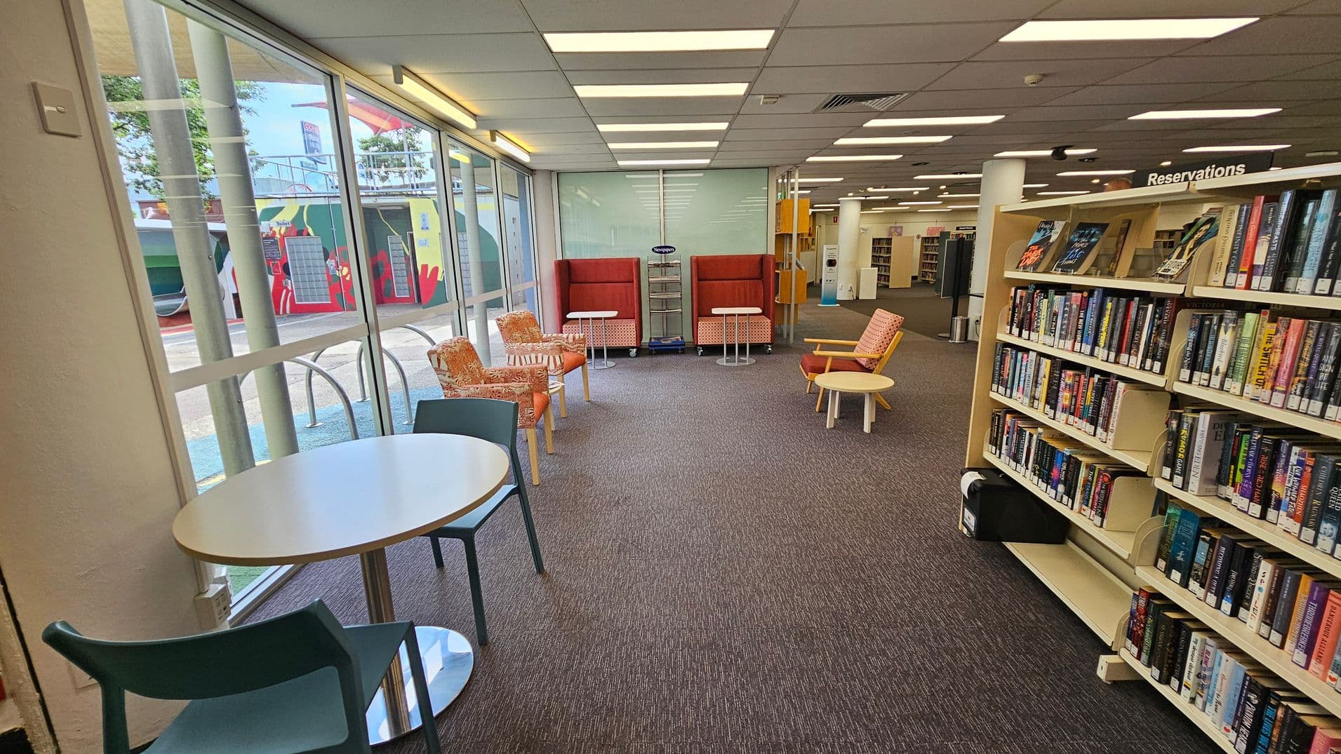 A library interior with a carpeted floor and a ceiling with fluorescent lights. On the left, large windows reveal an outdoor mural. In the centre, there are orange and patterned chairs with small round tables. On the right, a bookshelf labelled "Reservations" is filled with books. A small round table with a chair is near the window. The space is open and well-lit.