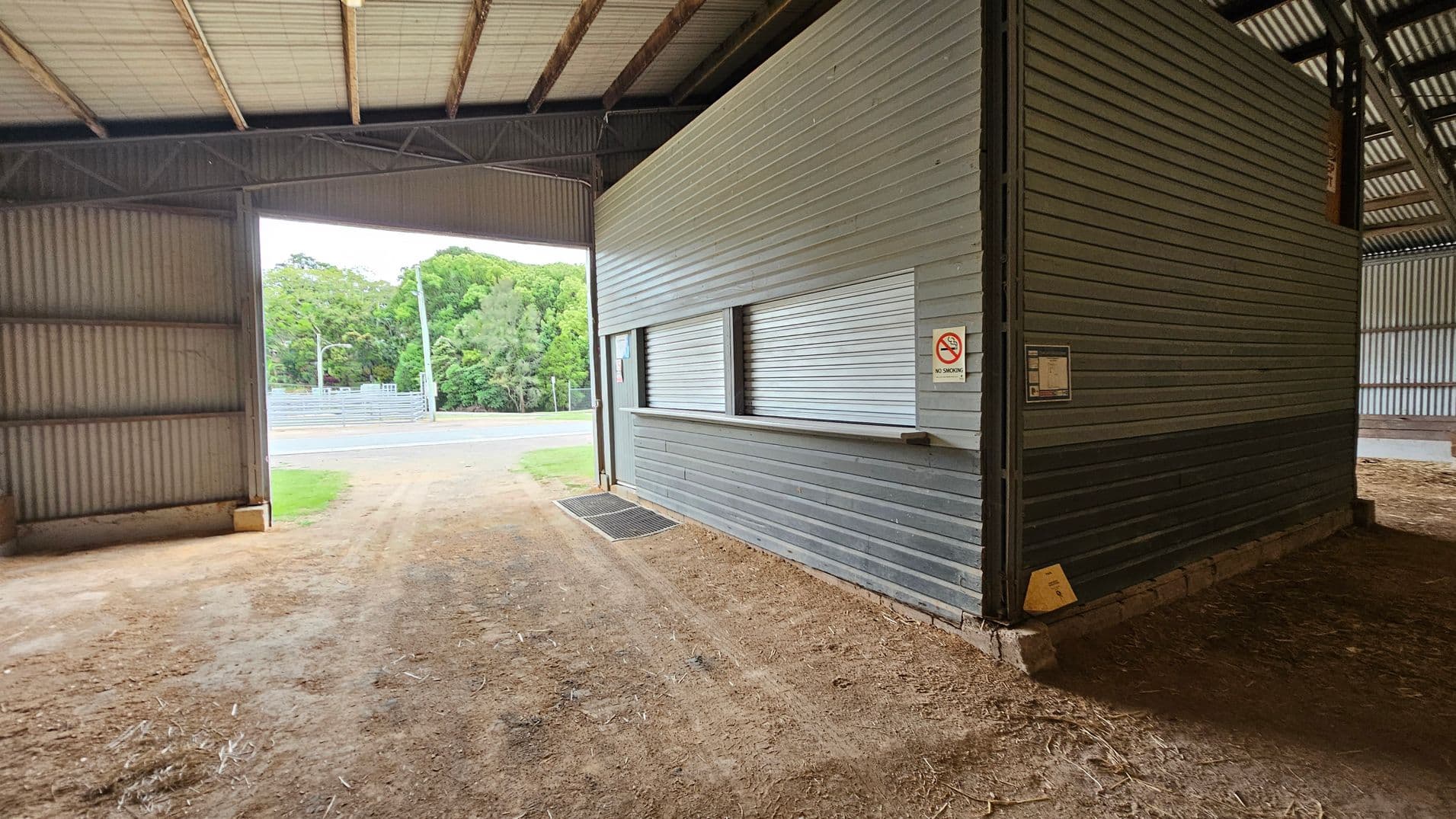 A large grey metal building labelled "Fergus Scott Pavilion" and "Dairy Cattle" is in the centre. It has two large open entrances. To the left, there's a smaller grey shed. The ground in front is a mix of gravel and grass. On the right, part of another structure with a sloped roof is visible. Trees are in the background on the left.