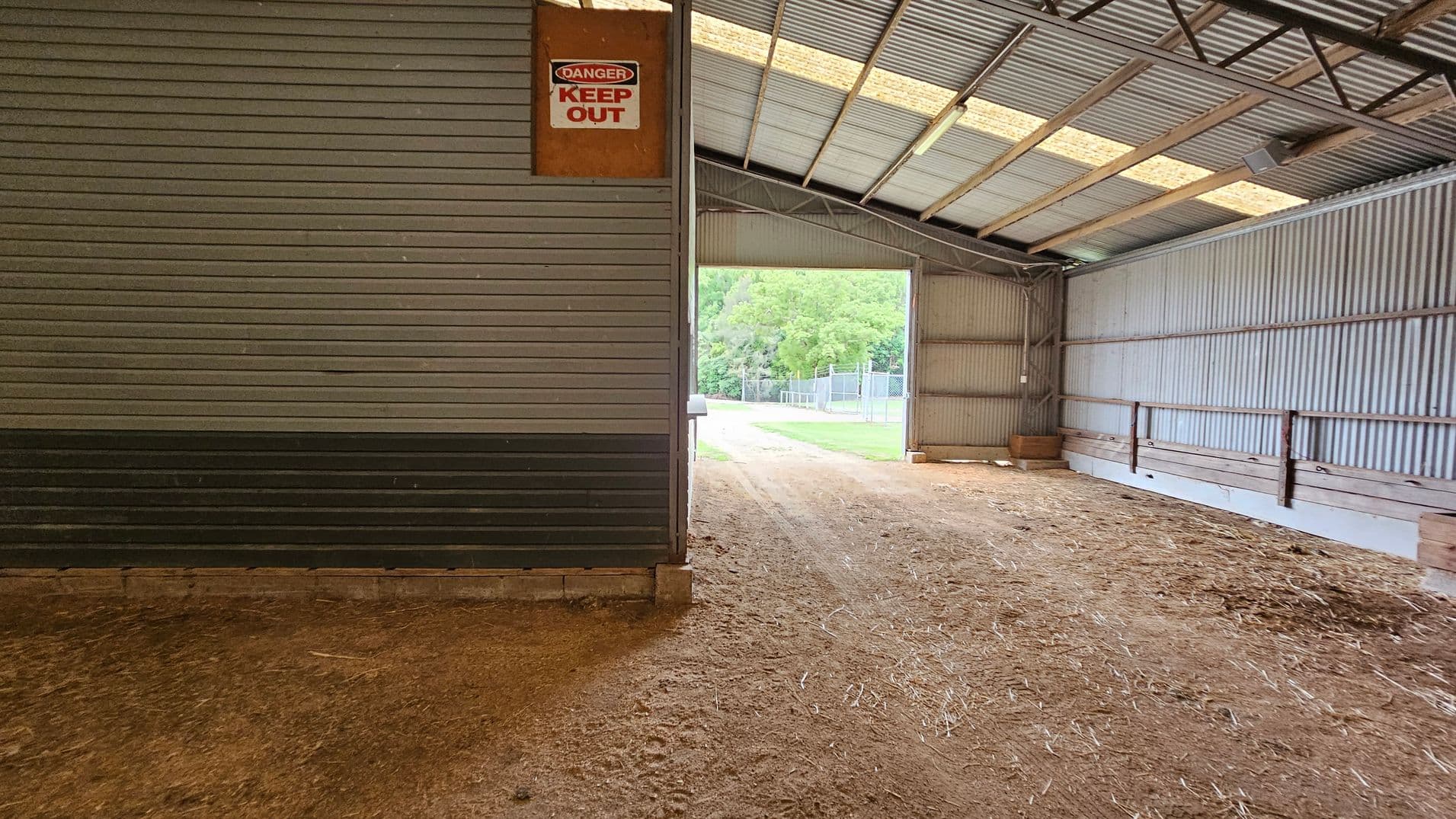 Inside a large, open-sided shed with a corrugated metal roof and walls. On the right, there's a small structure with horizontal cladding and closed shutters, featuring a "No Smoking" sign. The floor is dirt, with some grass and debris. Outside, visible through the open side, is a road bordered by green trees and a metal fence. The ceiling is high with exposed beams.