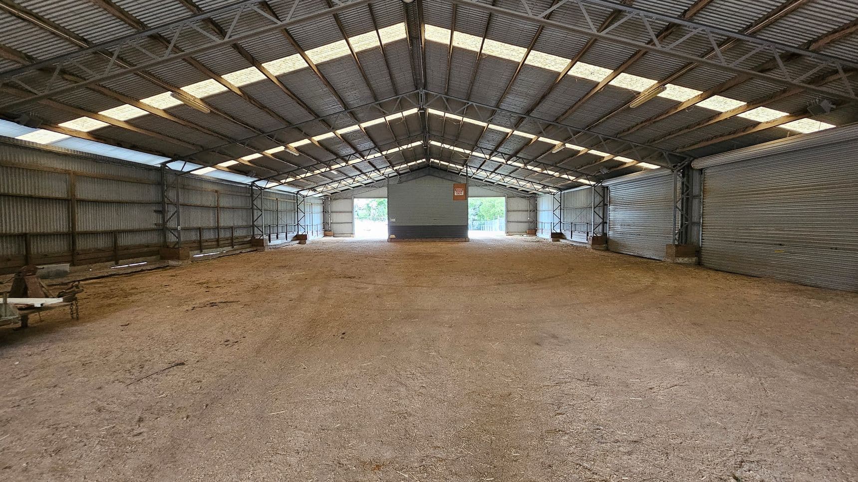 A large, empty warehouse with a corrugated metal roof and walls. The floor is covered with a mix of dirt and gravel. The ceiling has translucent panels allowing natural light to filter in. On the far end, there are two large open doorways with views of greenery outside. The interior is supported by metal beams, and there are some wooden pallets and equipment on the left side. The space is expansive and industrial in appearance.