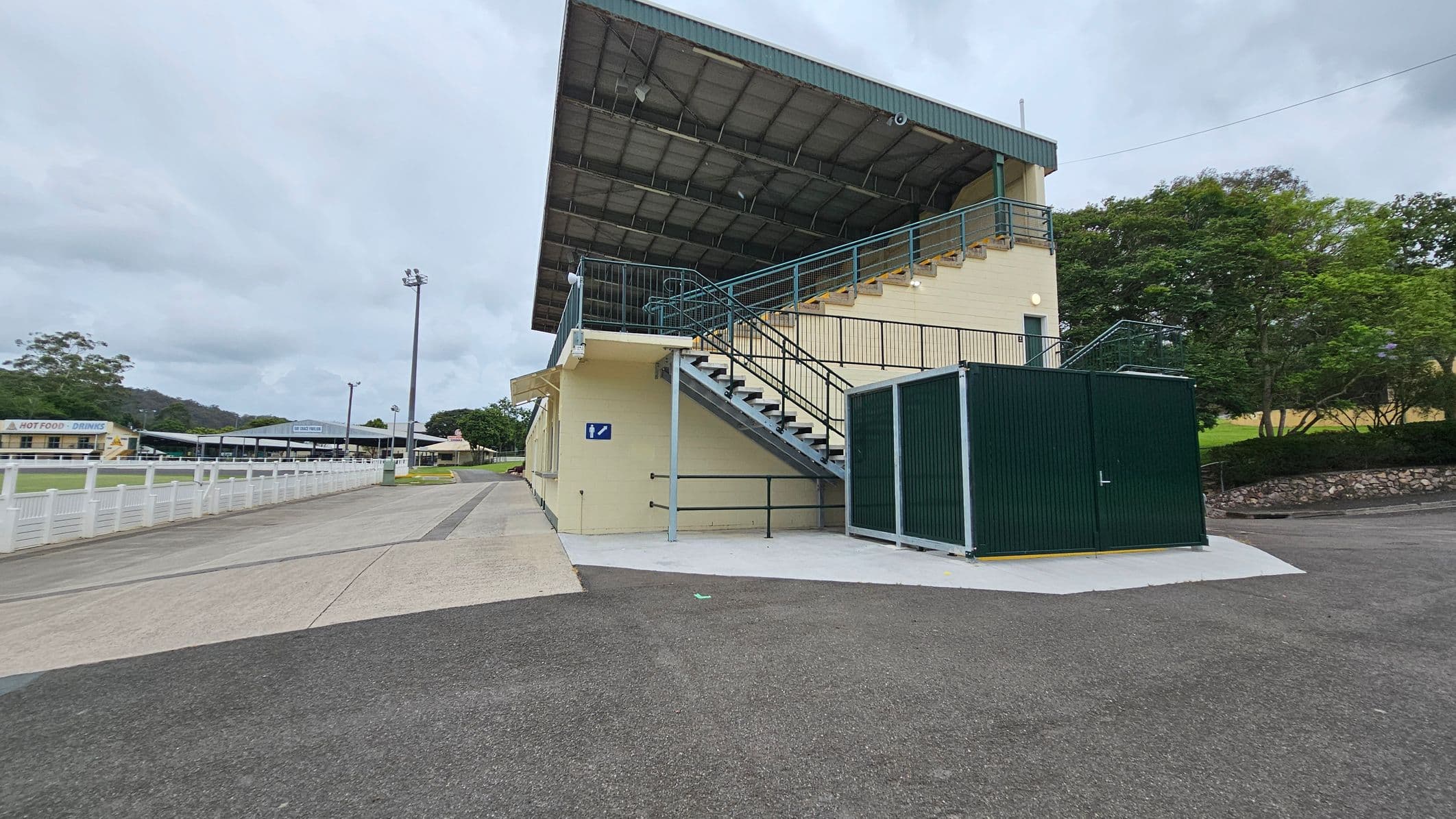 A sports facility with a covered grandstand on the right, featuring a row of green doors and large windows. The building is cream-coloured with green accents. The foreground shows a wide concrete pathway with patches of moss or lichen. In the background, there are tall floodlights and trees, with a smaller building visible to the left. The area appears open and spacious.