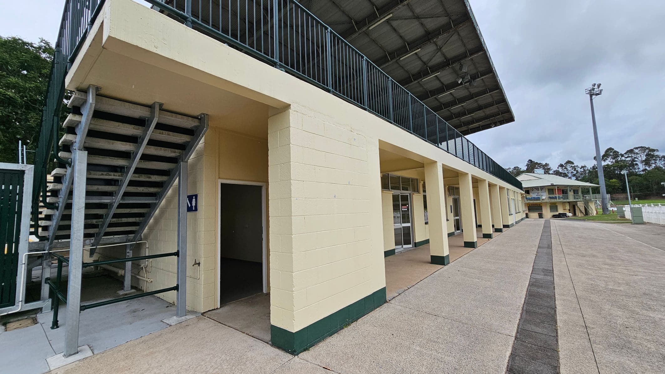 A sports facility with a two-storey structure. On the left, there is a staircase leading to the upper level, with a sign indicating a restroom below. The building has a series of columns and glass doors along the ground floor. The upper level features a railing and a covered roof. In the background, there is another similar building and a tall floodlight. The ground is paved with concrete, and there are trees in the distance.