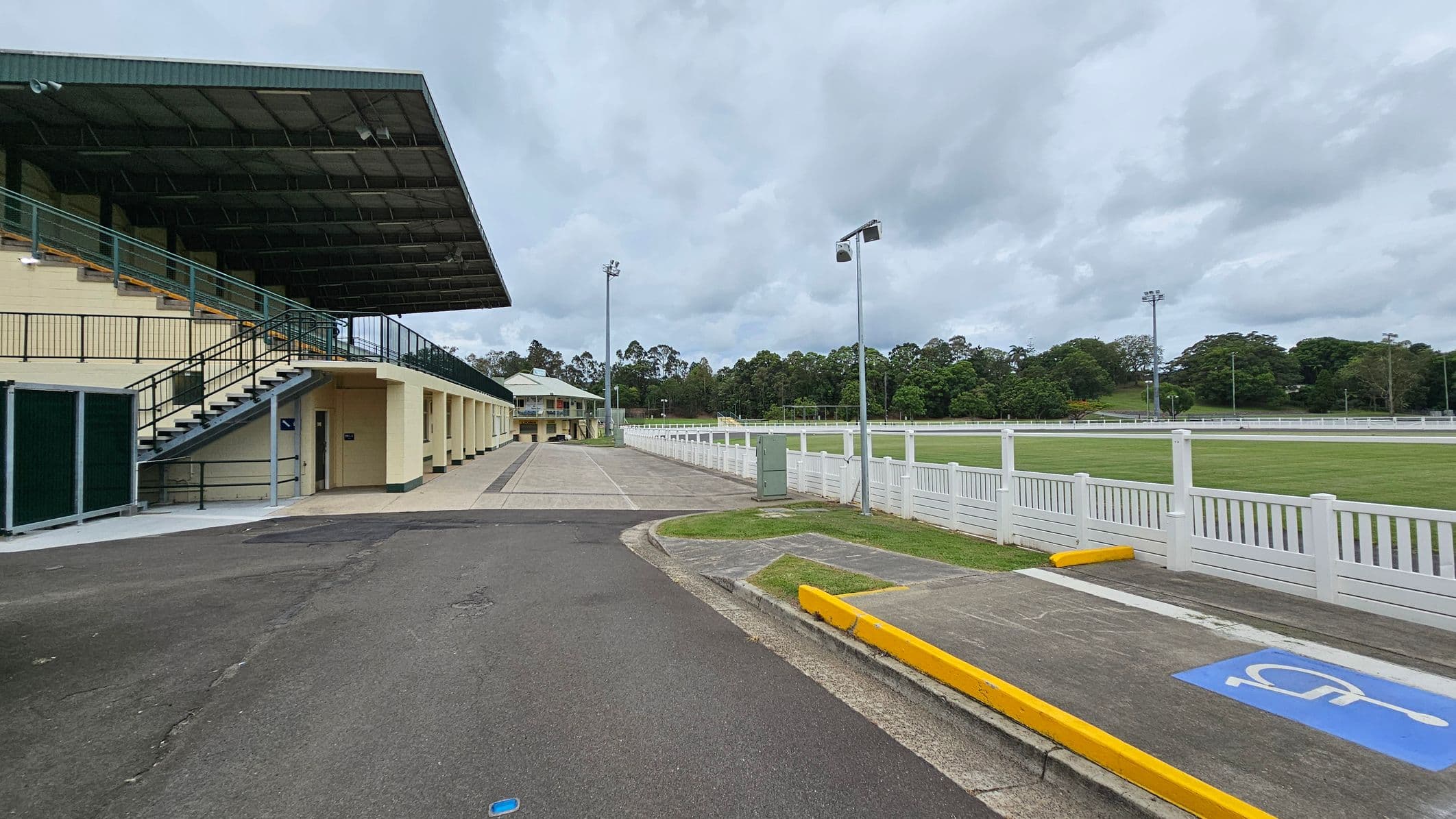 A sports facility with a covered grandstand on the left, featuring a staircase and multiple entrances. The ground is paved with a mix of asphalt and concrete. On the right, a white fence borders a grassy field. In the background, there are tall floodlights and a line of trees. A marked disabled parking space with a yellow kerb is visible in the foreground.