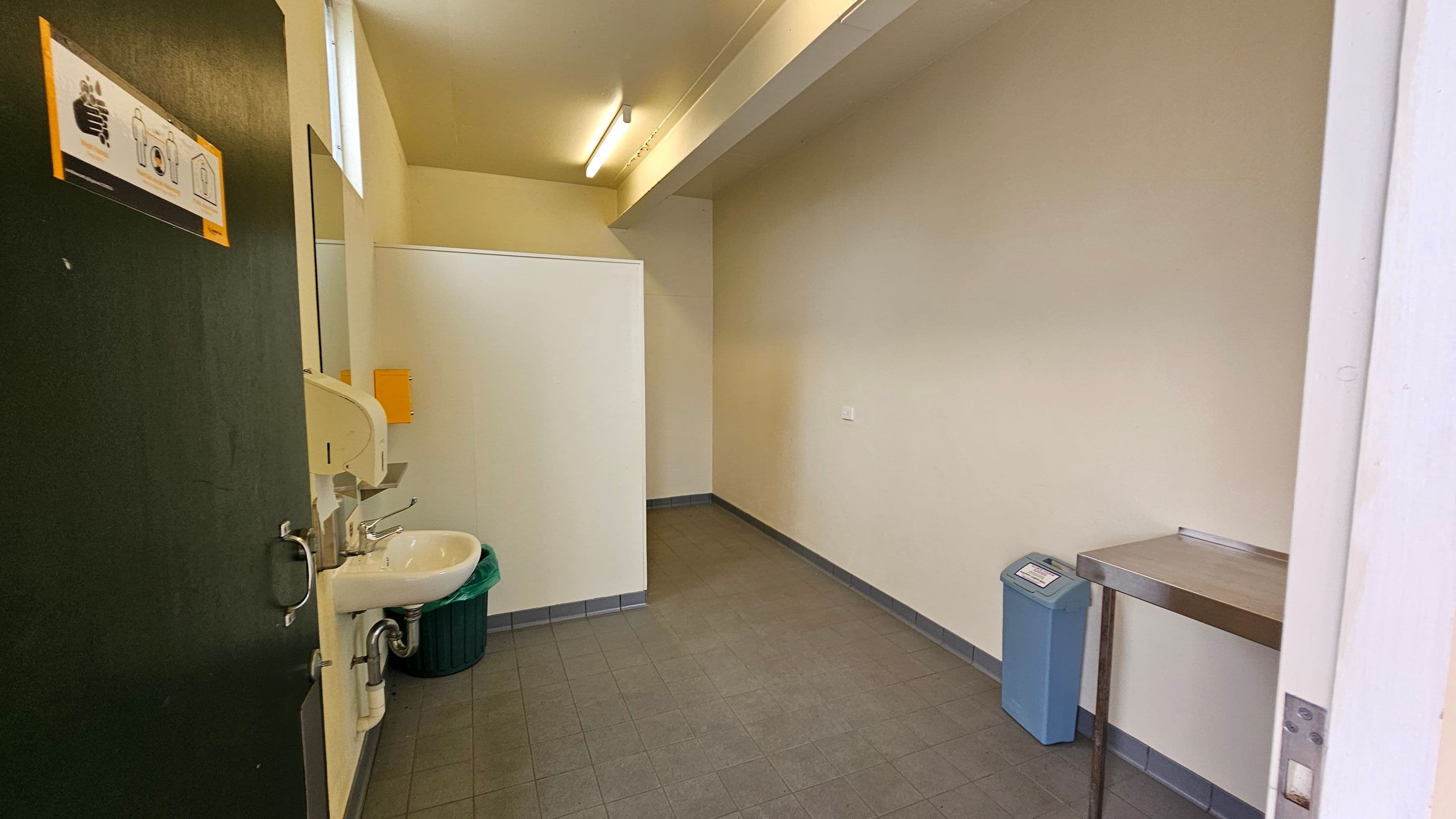A small room with a tiled floor and a light-coloured ceiling. On the left, there's a partially open door with a handwashing instruction sign. Next to it is a white sink with a soap dispenser and a green bin underneath. On the right, a metal table is attached to the wall, and a blue bin is nearby. The back wall is plain, with a small section of a partition visible. The room is lit by a fluorescent light.