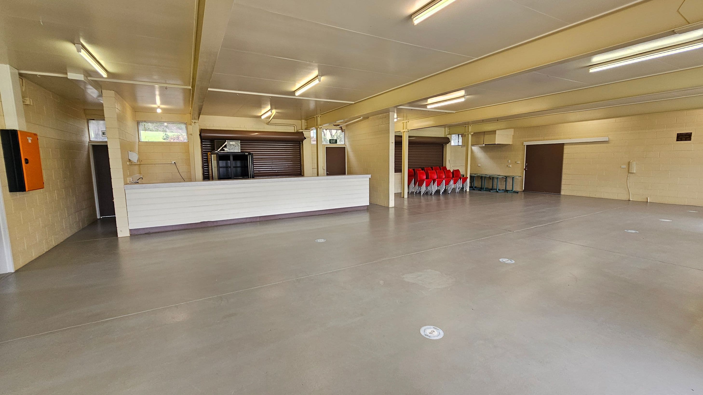 A spacious indoor area with a smooth, light brown floor and a white ceiling with fluorescent lights. On the left, there's a white counter with a small sink and an orange wall-mounted box. Behind the counter are two closed roller shutters. On the right, a stack of red chairs is neatly arranged against the wall. There are several doors, including an exit door, and a few small windows near the ceiling. The walls are light-coloured brick.