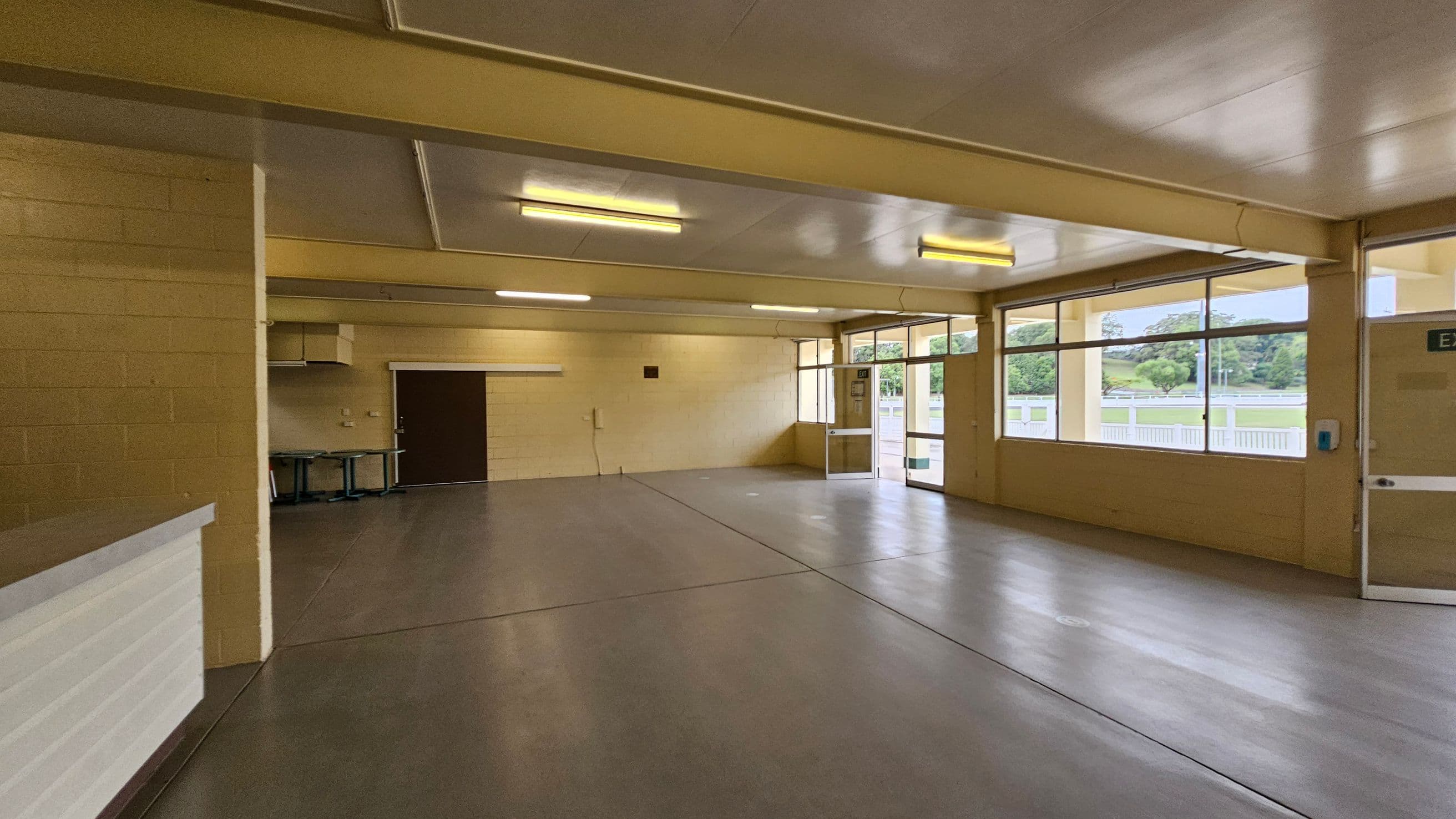 A spacious indoor room with a beige ceiling and floor. On the left, there's a white counter and a stack of red chairs against the wall. The back wall has a closed brown shutter and a door. On the right, large windows and a glass door provide natural light, with a view of an outdoor area. The room is lit by fluorescent lights on the ceiling.
