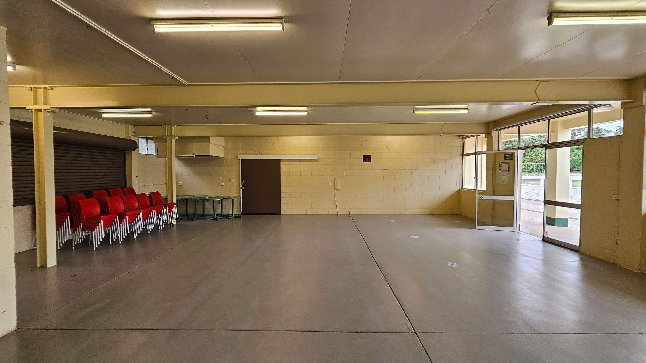 A spacious, empty room with beige walls and a smooth grey floor. On the left, there's a counter with a white top. The back wall features a dark brown door and a small kitchen area with a range hood. Large windows on the right let in natural light, showing a view of greenery outside. The ceiling is white with fluorescent lights. An exit door is on the far right.