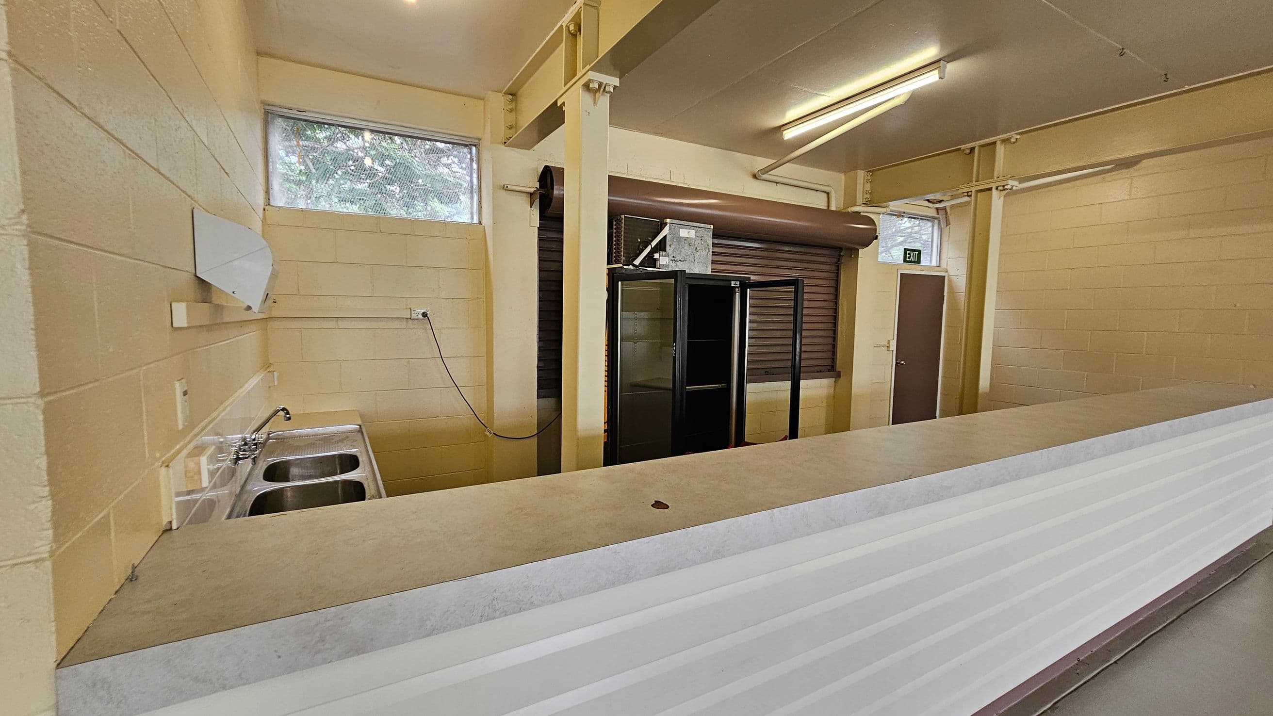 A room with cream-coloured brick walls and a long counter in the foreground. On the left, there's a double sink with a tap and a small window above it. In the middle, a black-framed glass cabinet stands next to a closed roller shutter. On the right, a brown door with an "EXIT" sign above it. The ceiling is fitted with fluorescent lights, and the floor is not visible.