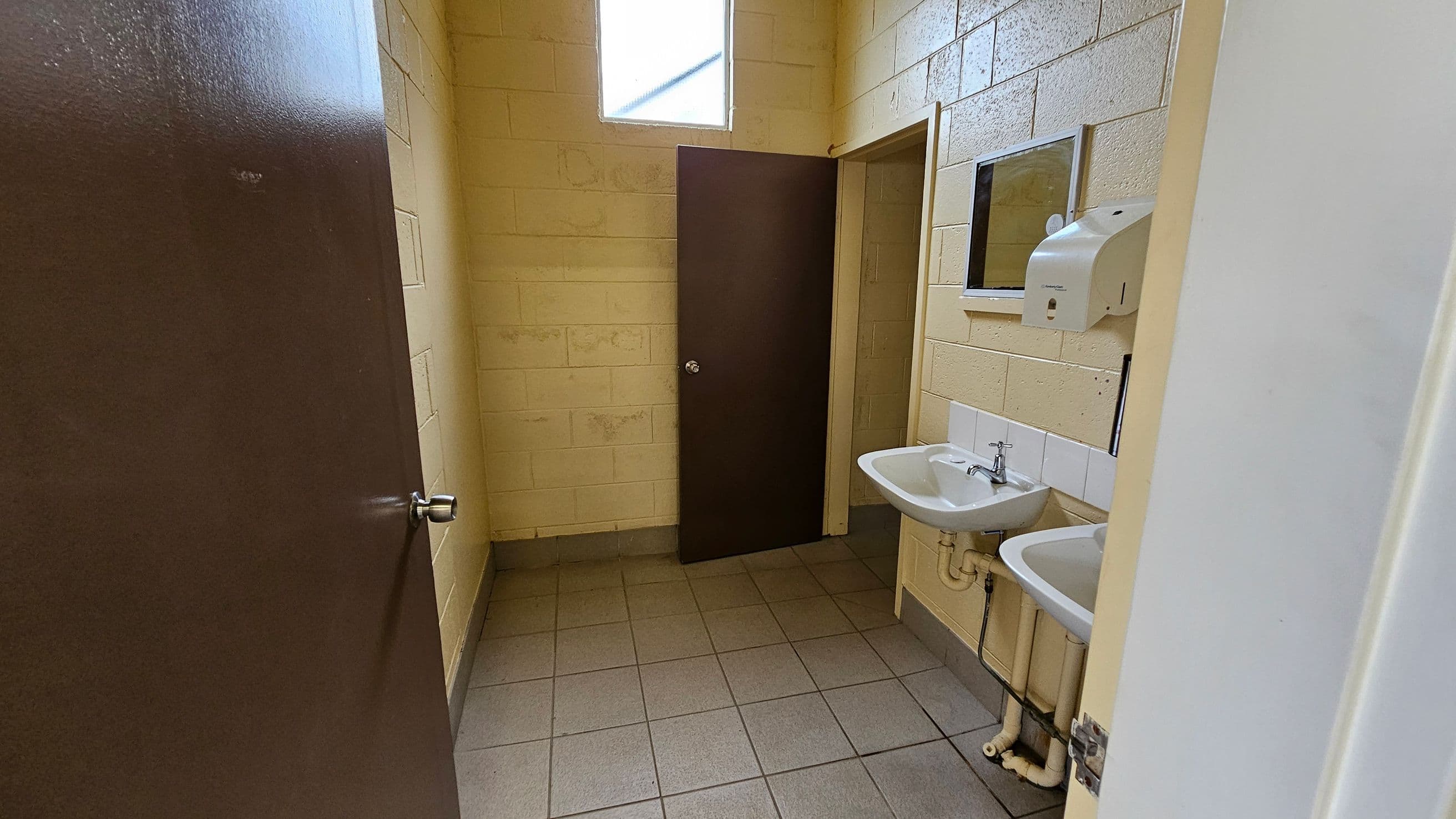 A small bathroom with beige brick walls and a tiled floor. On the right, there are two white sinks with a mirror above and a soap dispenser mounted on the wall. In the centre, a brown door is slightly ajar, leading to another room. Above the door, a high window allows natural light to enter. The ceiling is not visible.