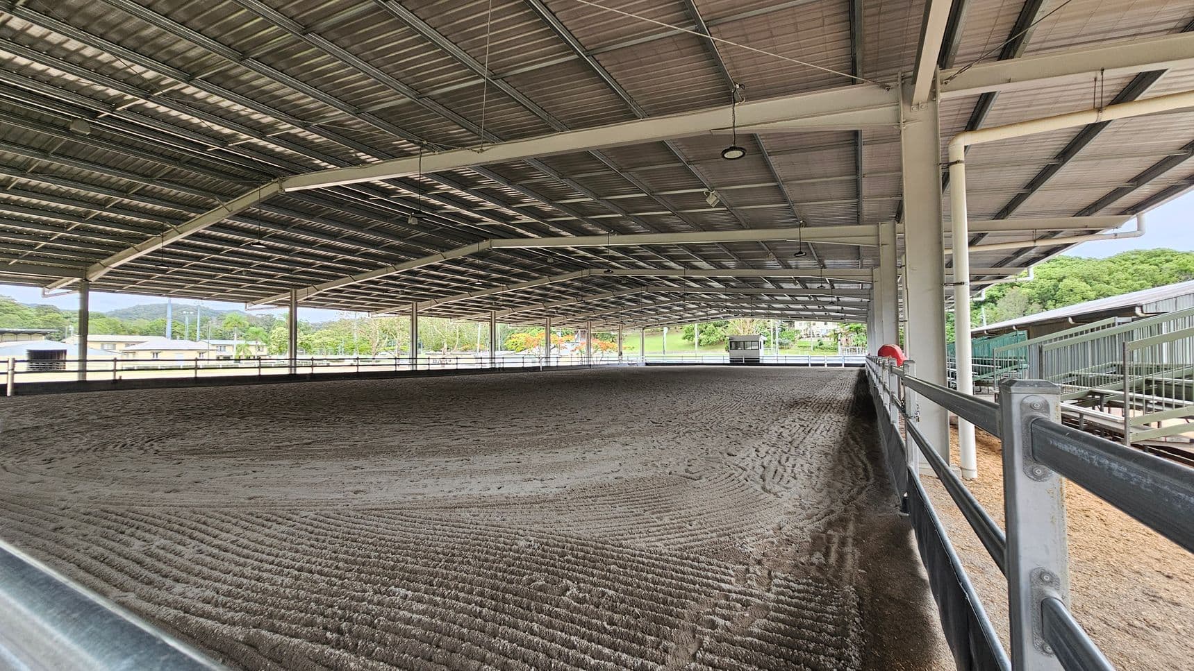 A large covered equestrian arena with a metal roof and open sides. The ground is textured with a mix of sand and soil. On the right, a metal railing runs along the perimeter. In the background, there are trees and some buildings visible through the open sides. The ceiling is supported by metal beams and has several lights hanging from it.
