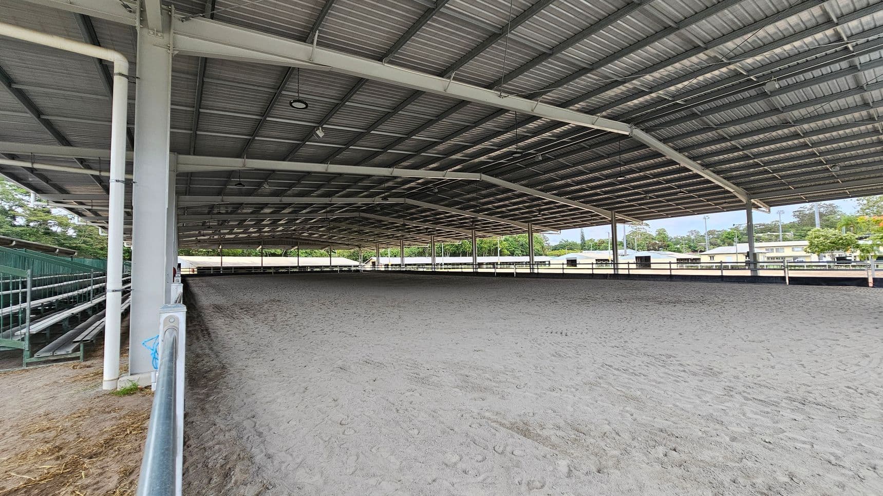 A large covered equestrian arena with a metal roof and open sides. The ground is sandy, showing hoof prints. On the left, there are green metal bleachers for spectators. The background features trees and some buildings visible through the open sides. The ceiling is supported by metal beams and has several lights hanging from it.