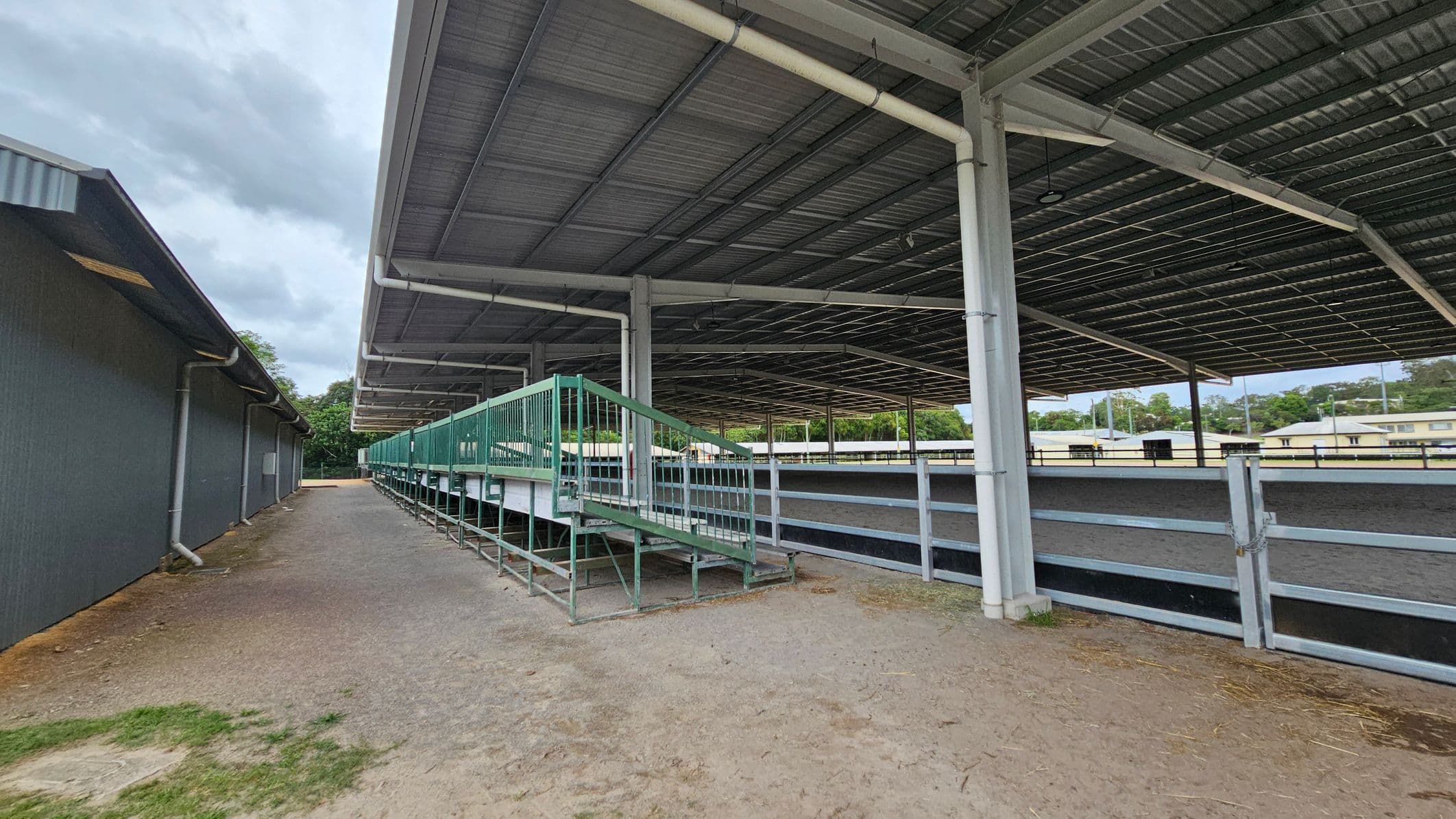 A covered outdoor area with a large metal roof supported by white pillars. On the right, there is a fenced area with a green metal ramp and seating. The ground is a mix of dirt and gravel. On the left, a long, dark building with a sloped roof runs parallel to the covered area. In the background, there are trees and some low buildings.