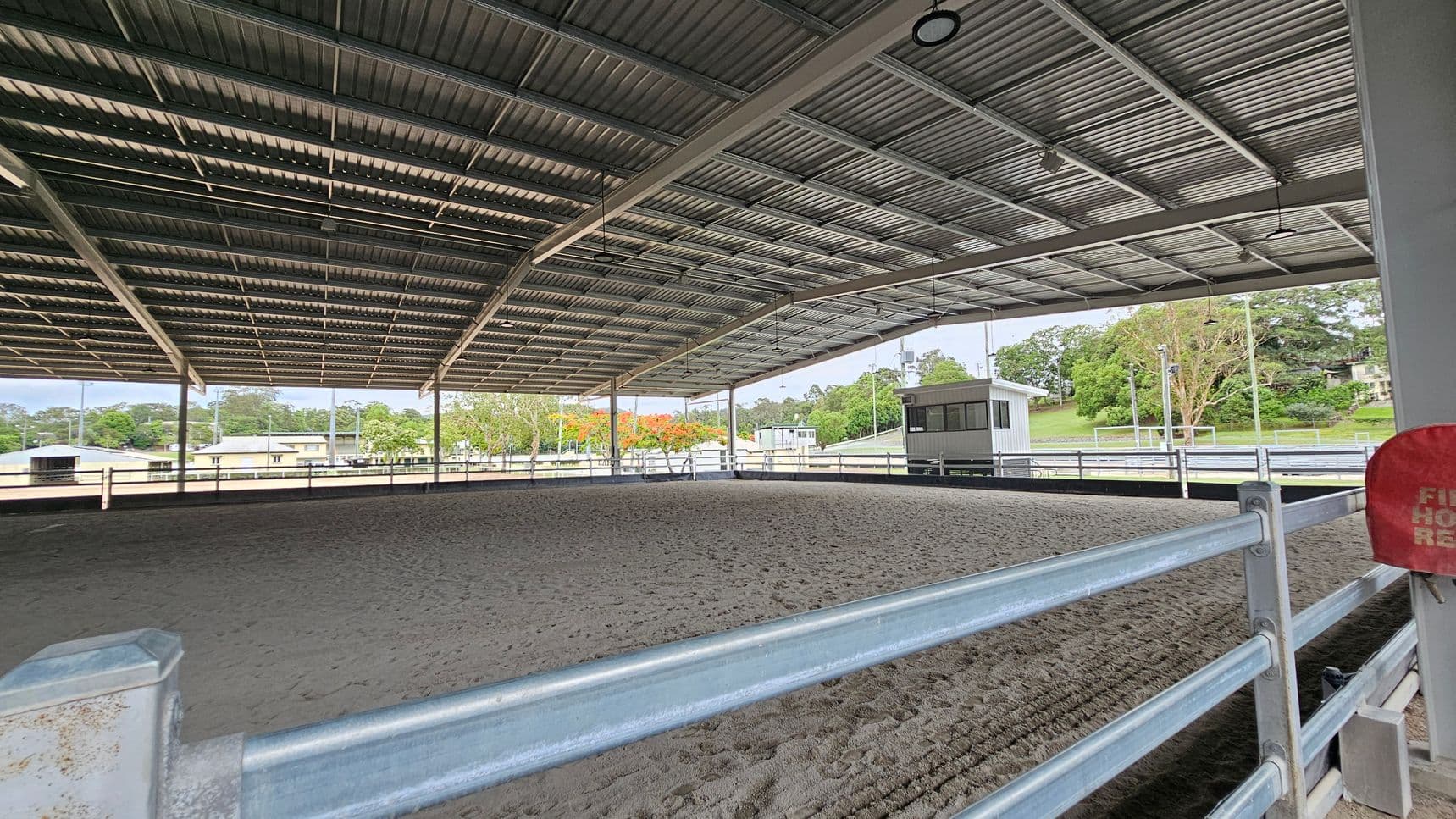 A covered equestrian arena with a metal roof and open sides. The ground is sandy, and a metal railing surrounds the area. In the background, there are trees and a small building. A red fire hose reel is visible on the right side. The ceiling has several lights and fans.
