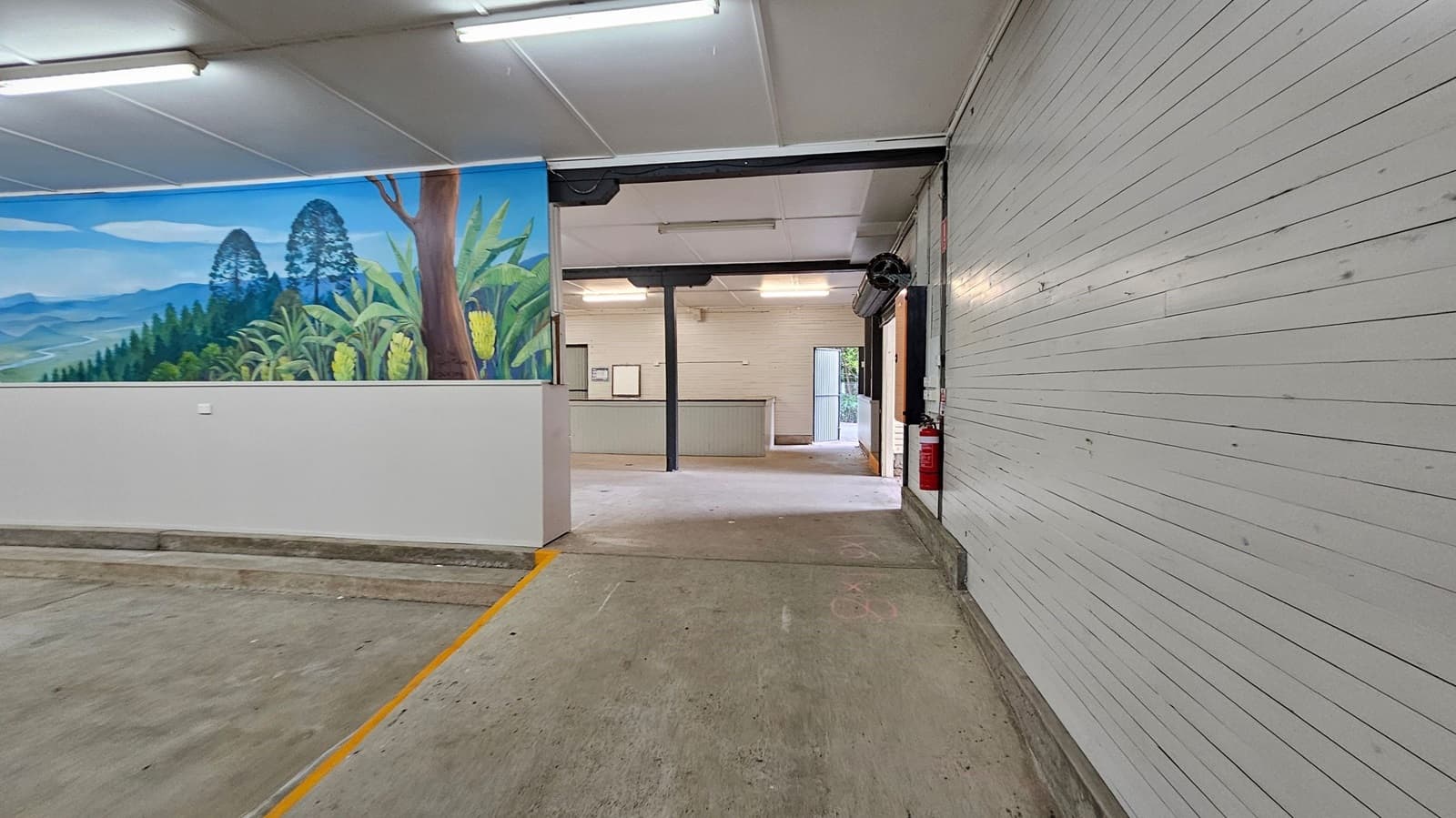 A room with a concrete floor and a white panelled ceiling. On the left, there's a mural depicting a landscape with trees and plants. In the centre, an open doorway leads to another room with a counter and a door to the outside. On the right, a white panelled wall with a fire extinguisher mounted near the doorway. Fluorescent lights are fixed to the ceiling.
