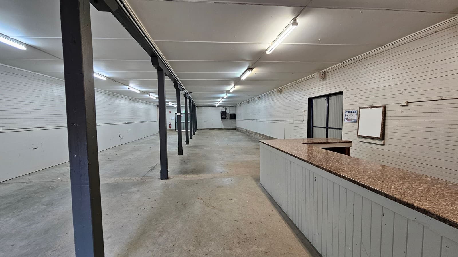 A long, empty indoor space with a concrete floor and a white panelled ceiling. The left and right walls are lined with white panels. A series of black metal support beams run down the centre. At the far end, there is a double door with a metal frame and a fire extinguisher nearby. On the right, there is a small partitioned area with a notice board and a door. The lighting is provided by fluorescent tubes on the ceiling.